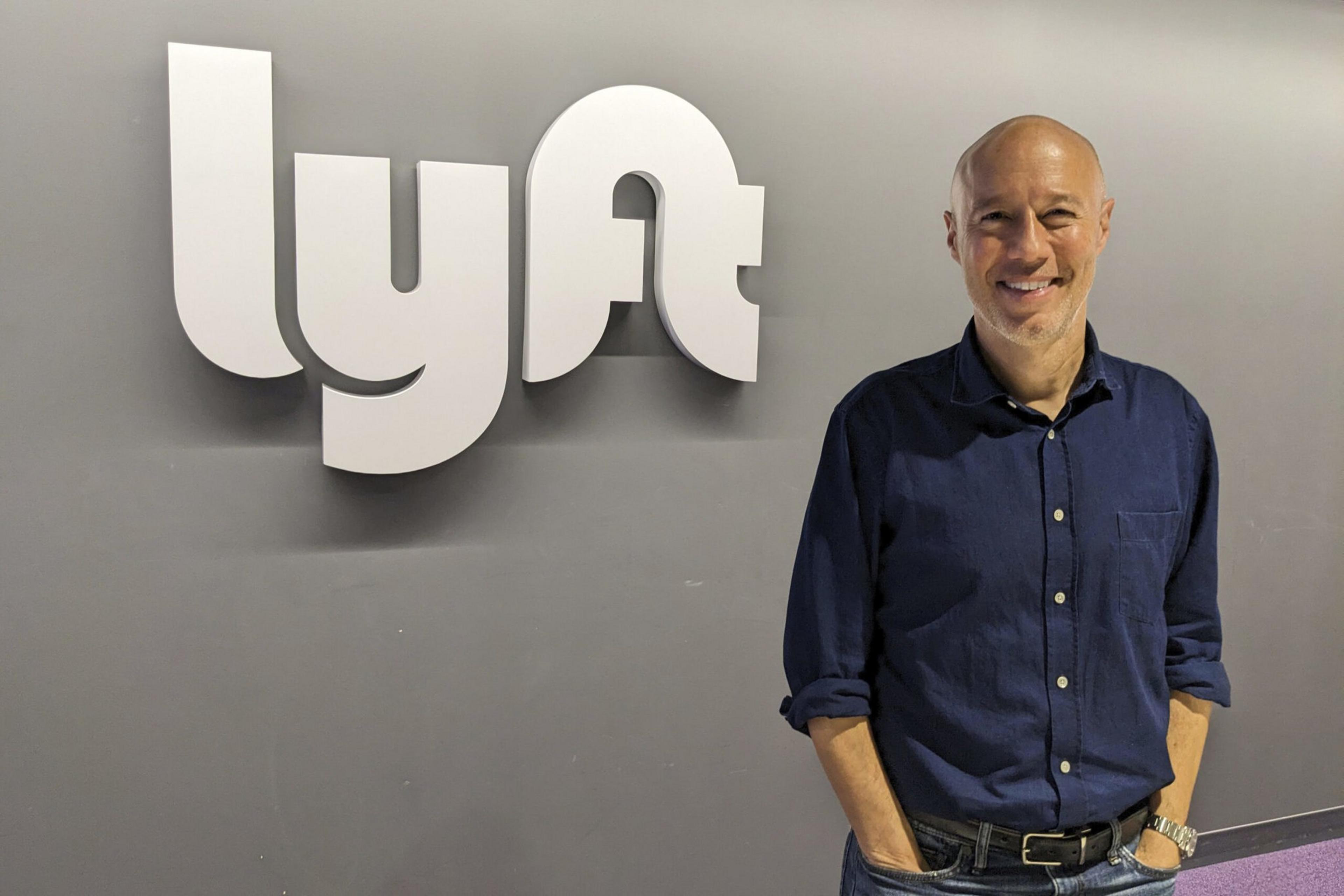 A bald man wearing a dark blue button-up shirt and jeans smiles while standing with hands in pockets next to a large white "lyft" logo on a gray wall.