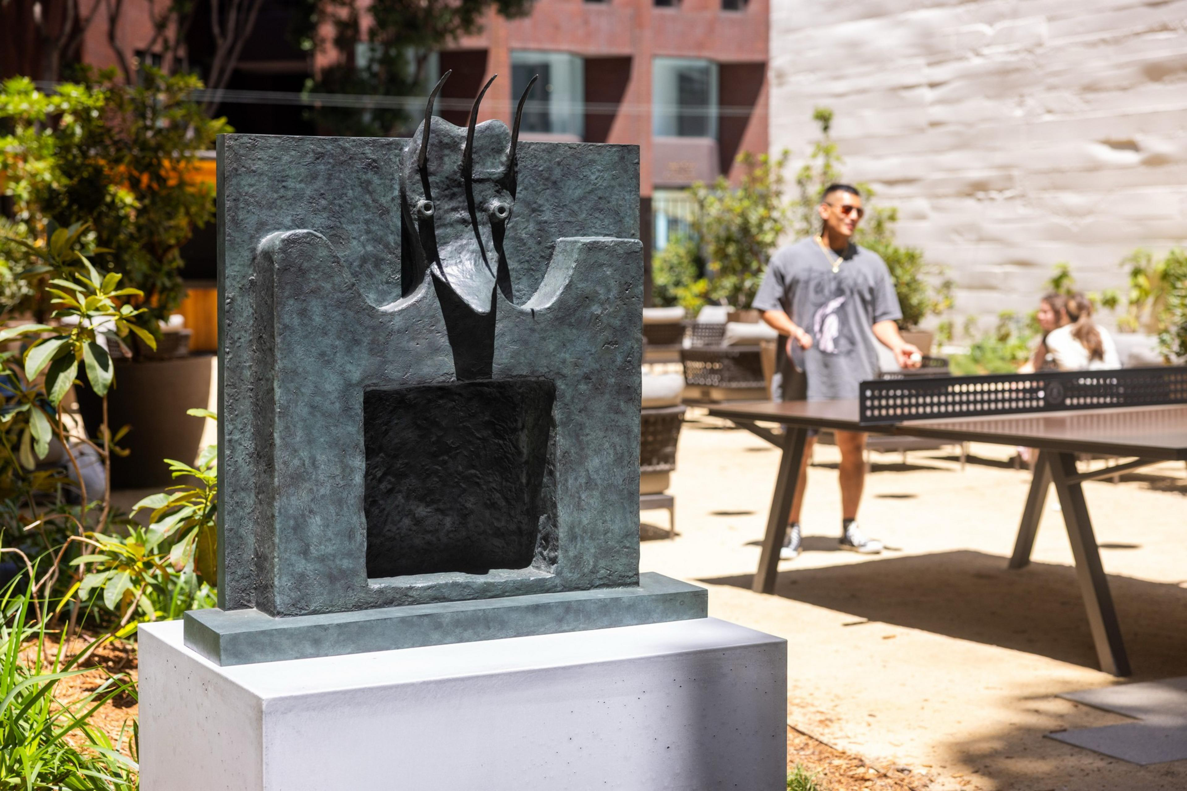 A modern sculpture with angular shapes and pointed features stands outdoors. In the background, a person plays table tennis under sunlight.