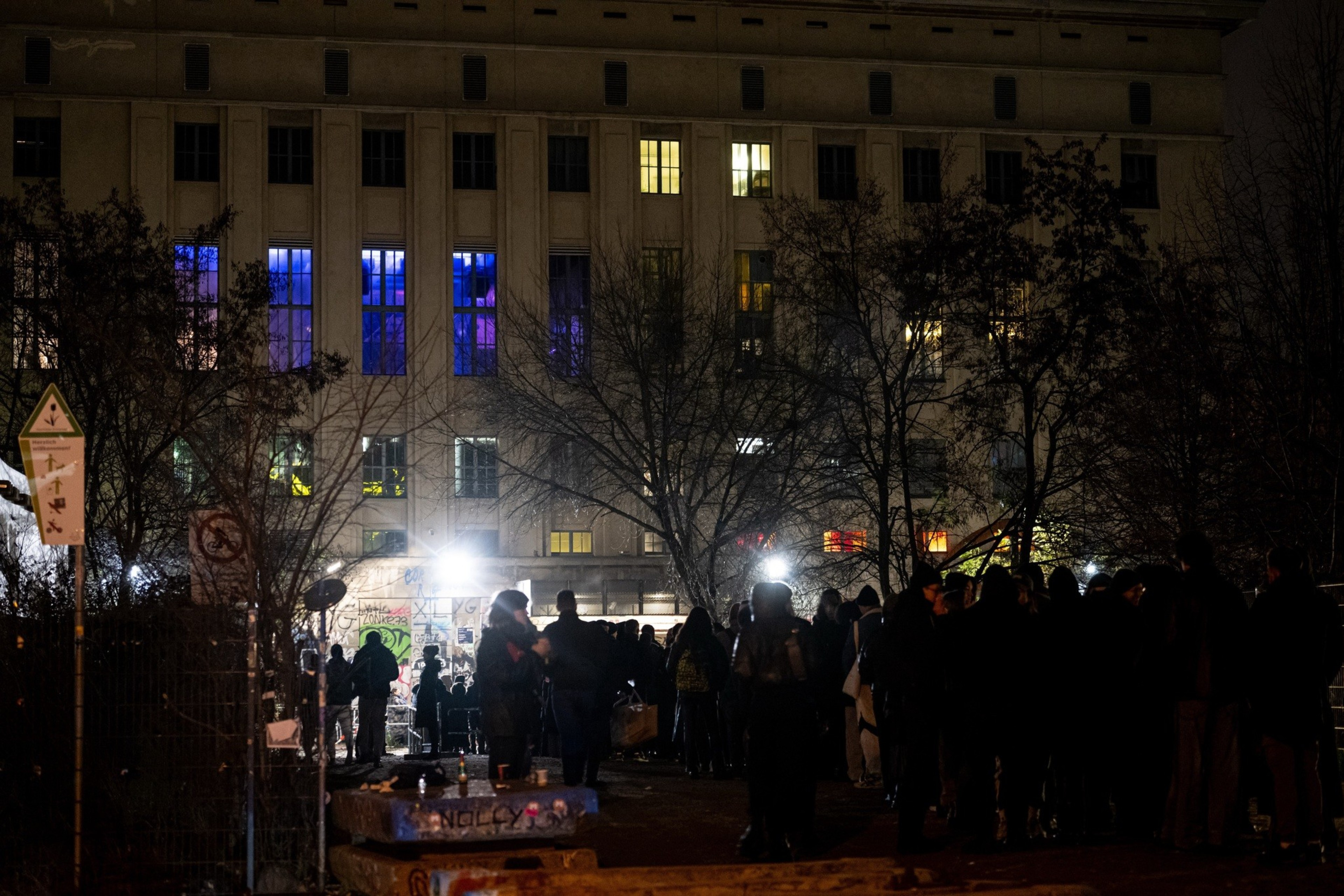 A crowd of people gathers outside a building at night, illuminated by bright lights and colorful windows in the background.