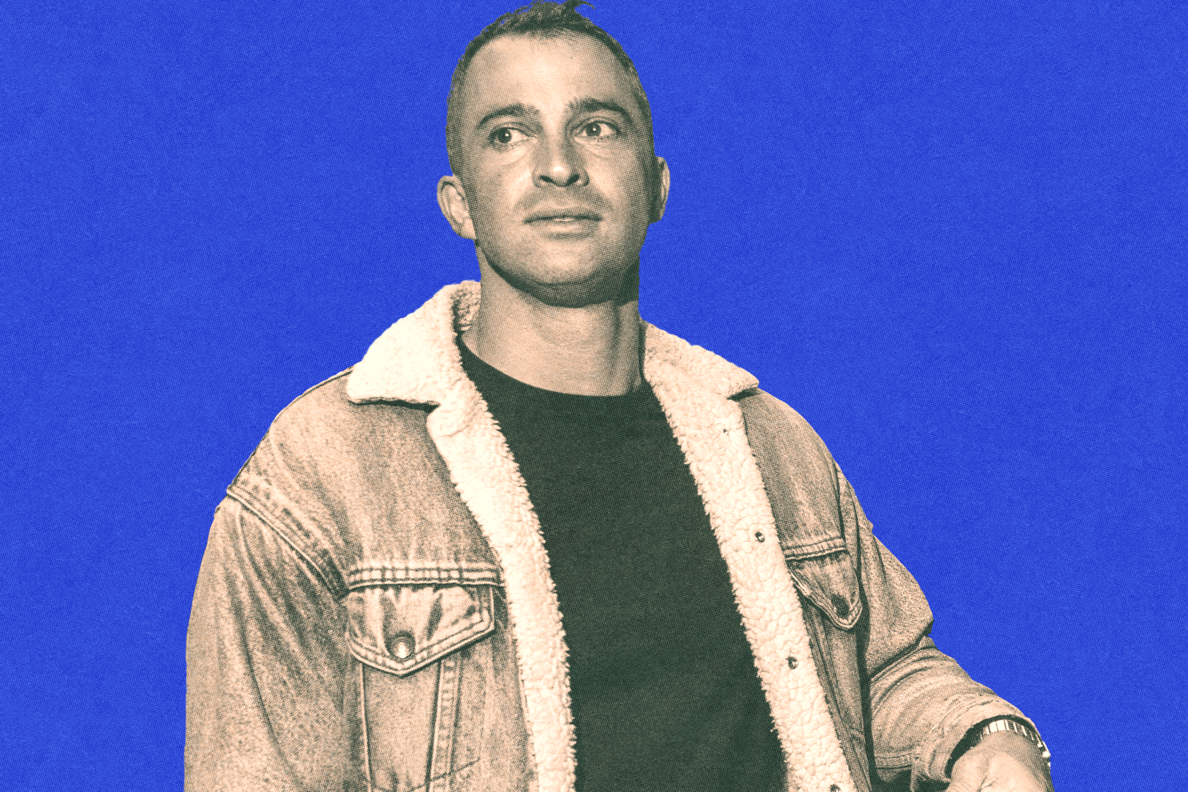 A man wearing a shearling-lined denim jacket and a black shirt looks slightly to the side against a solid blue background.