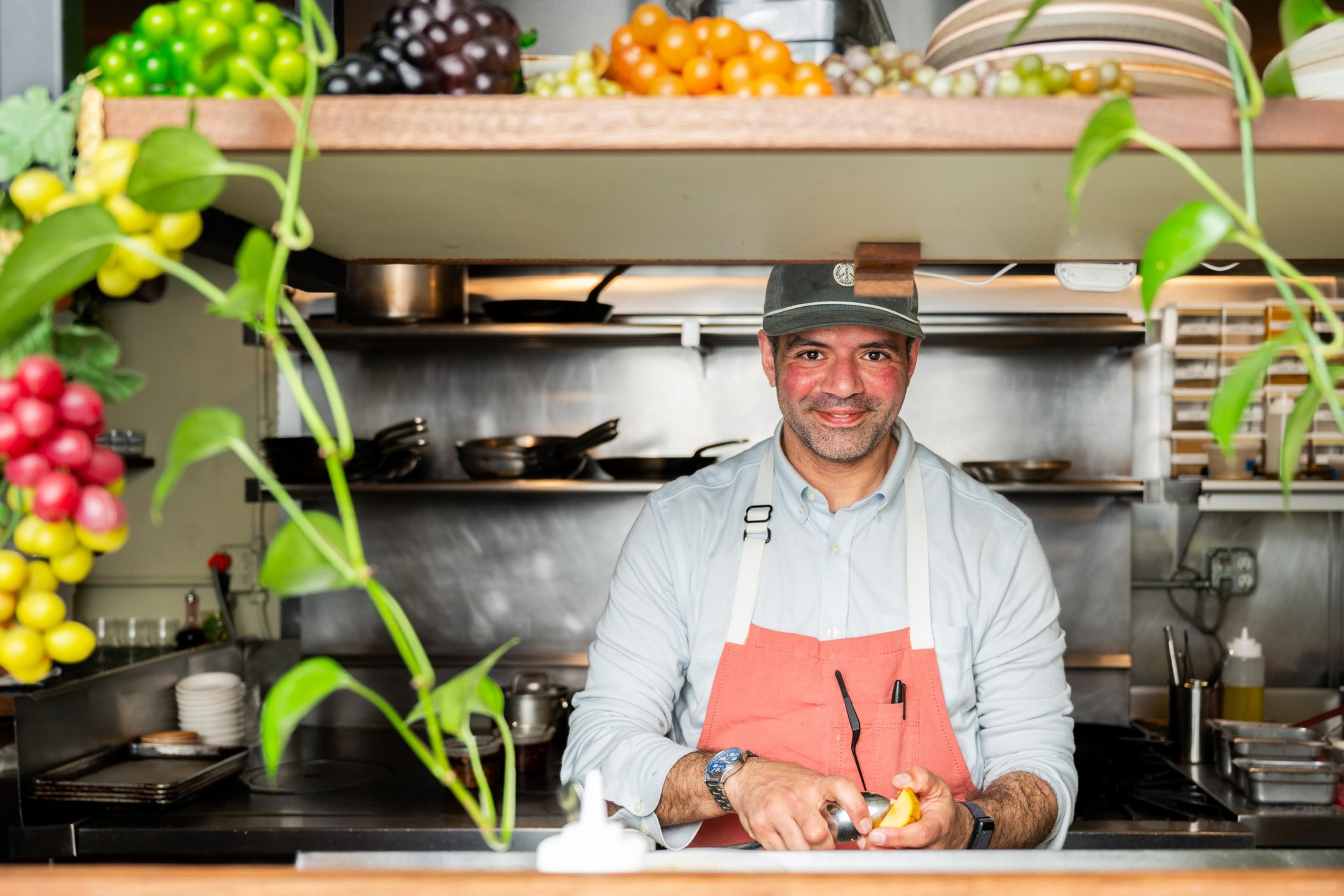 A smiling person in an apron peels an orange at a kitchen counter, with shelves of cookware and fresh fruit above. Green plants add a touch of color.