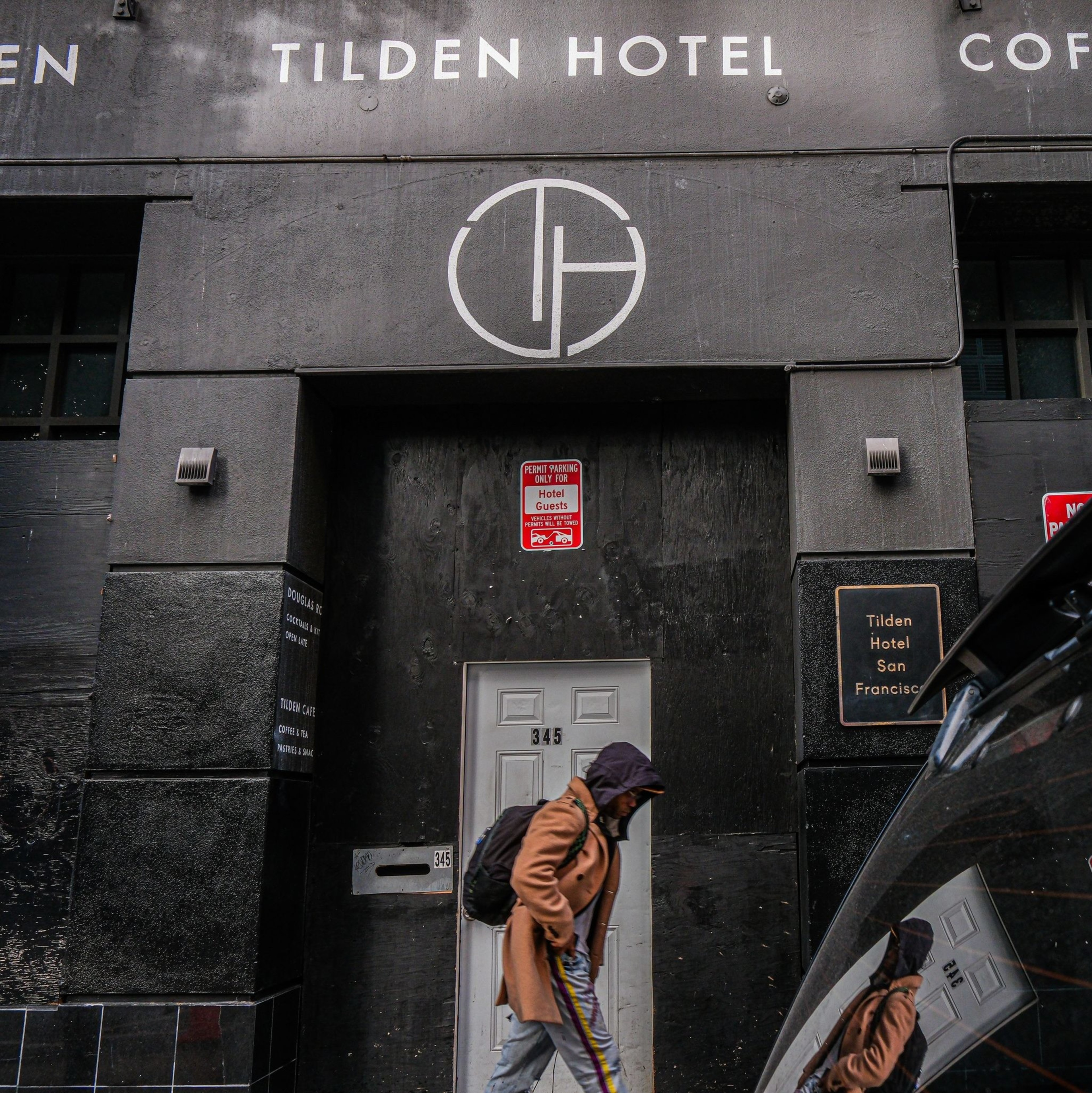 A person in a coat and hood walks past the entrance of Tilden Hotel. The building's facade is dark with signage and a distinct round logo above the door.