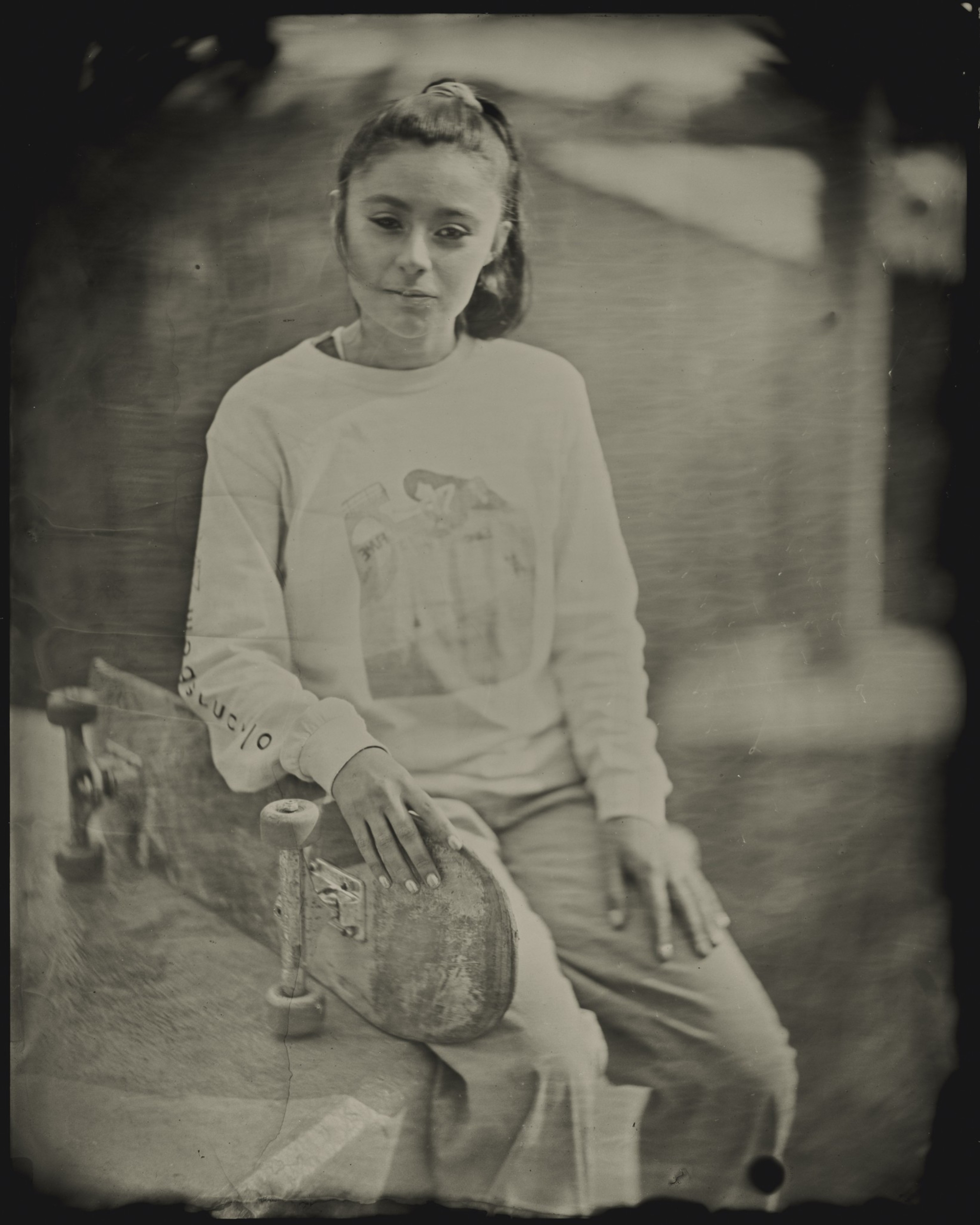 A person with long hair sits holding a skateboard. They're wearing a sweater with a graphic design. The image has a vintage, sepia tone.