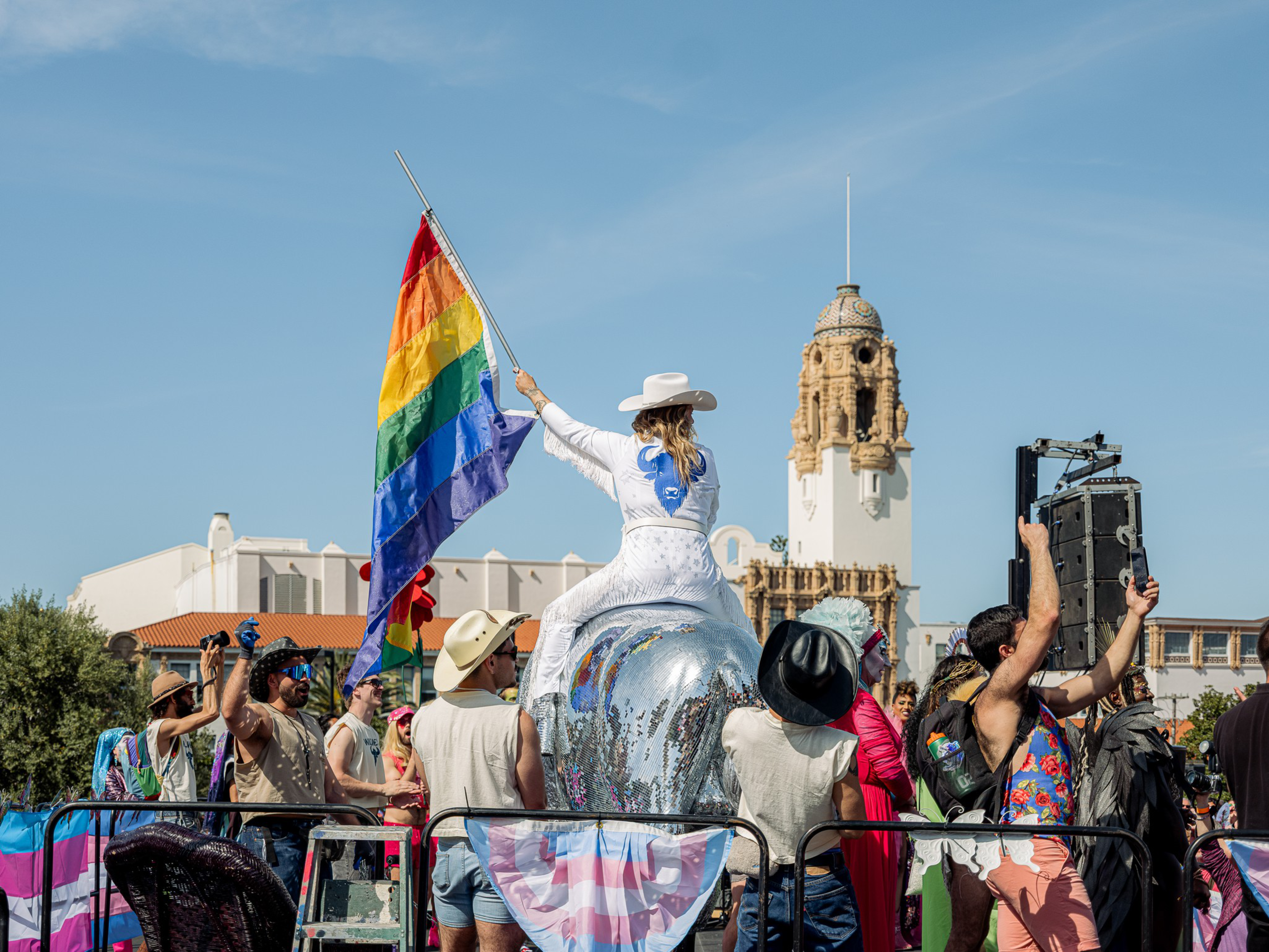A person in a white outfit and hat rides a shiny float with a rainbow flag, surrounded by people in festive attire, against a blue sky and a historic building.