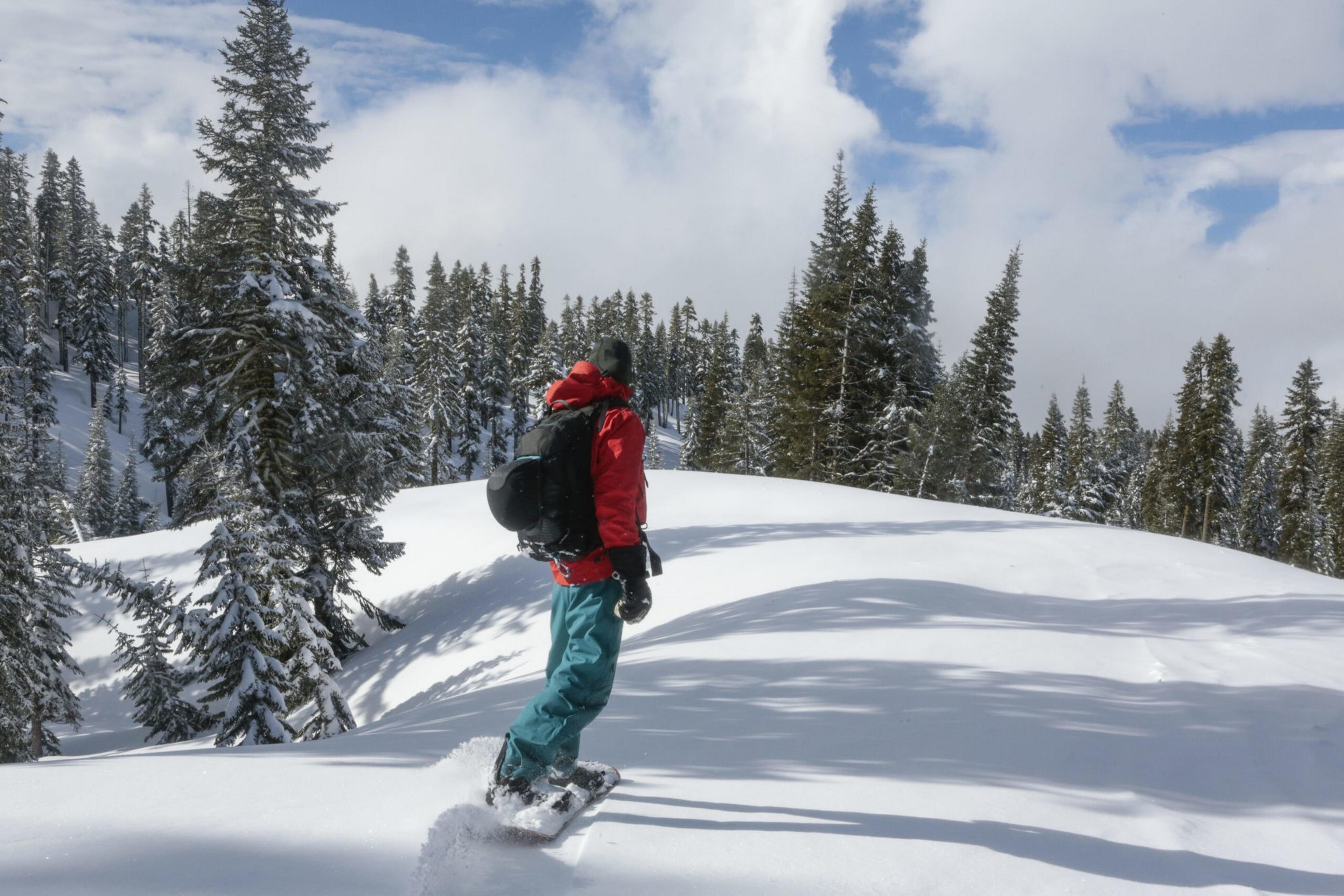 A person wearing a red jacket and teal pants snowboards through deep snow surrounded by snow-covered pine trees under a partly cloudy sky.