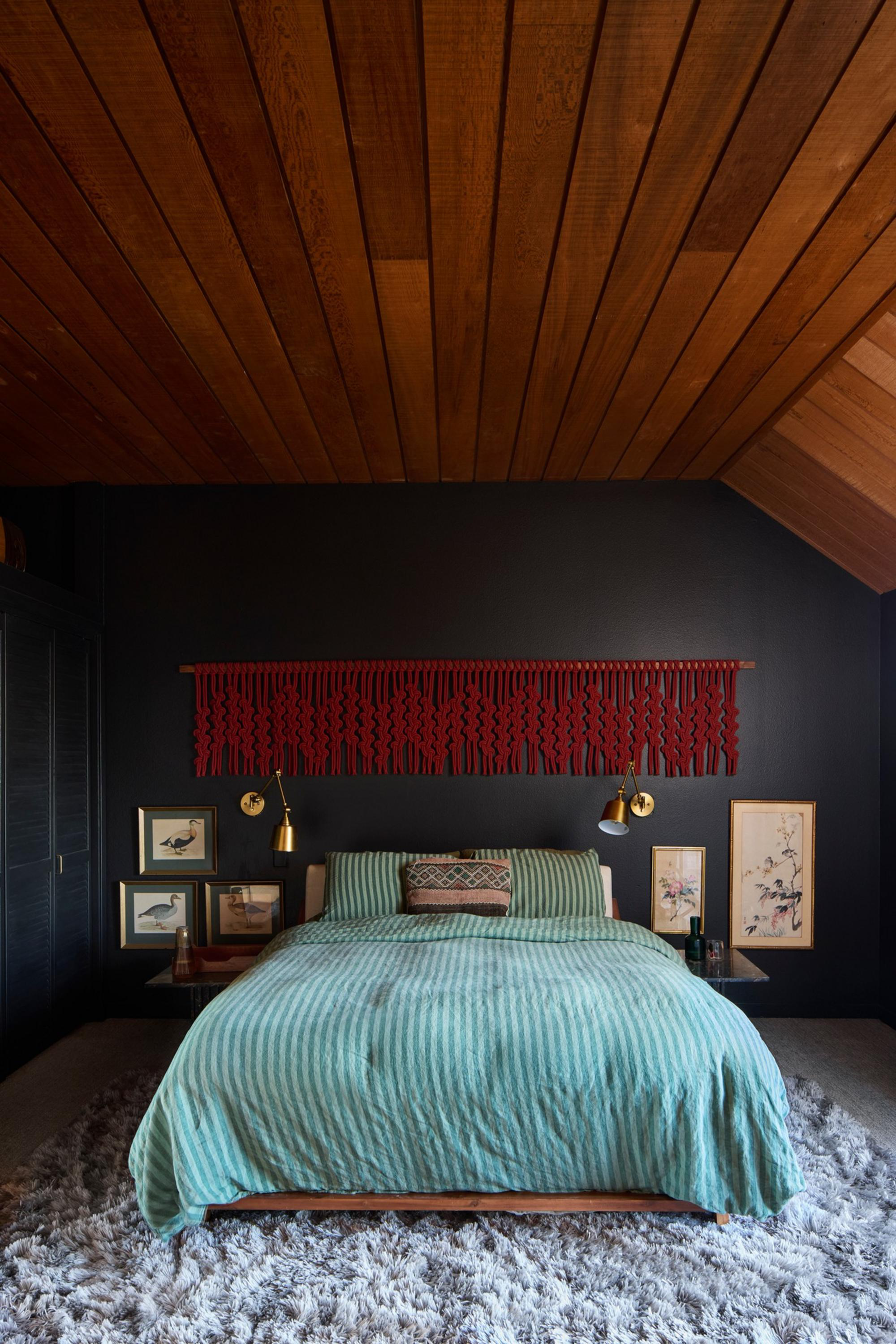 A bed with green striped bedding sits against a dark wall, flanked by framed bird and floral art, with gold wall lamps and a red macramé hanging above.