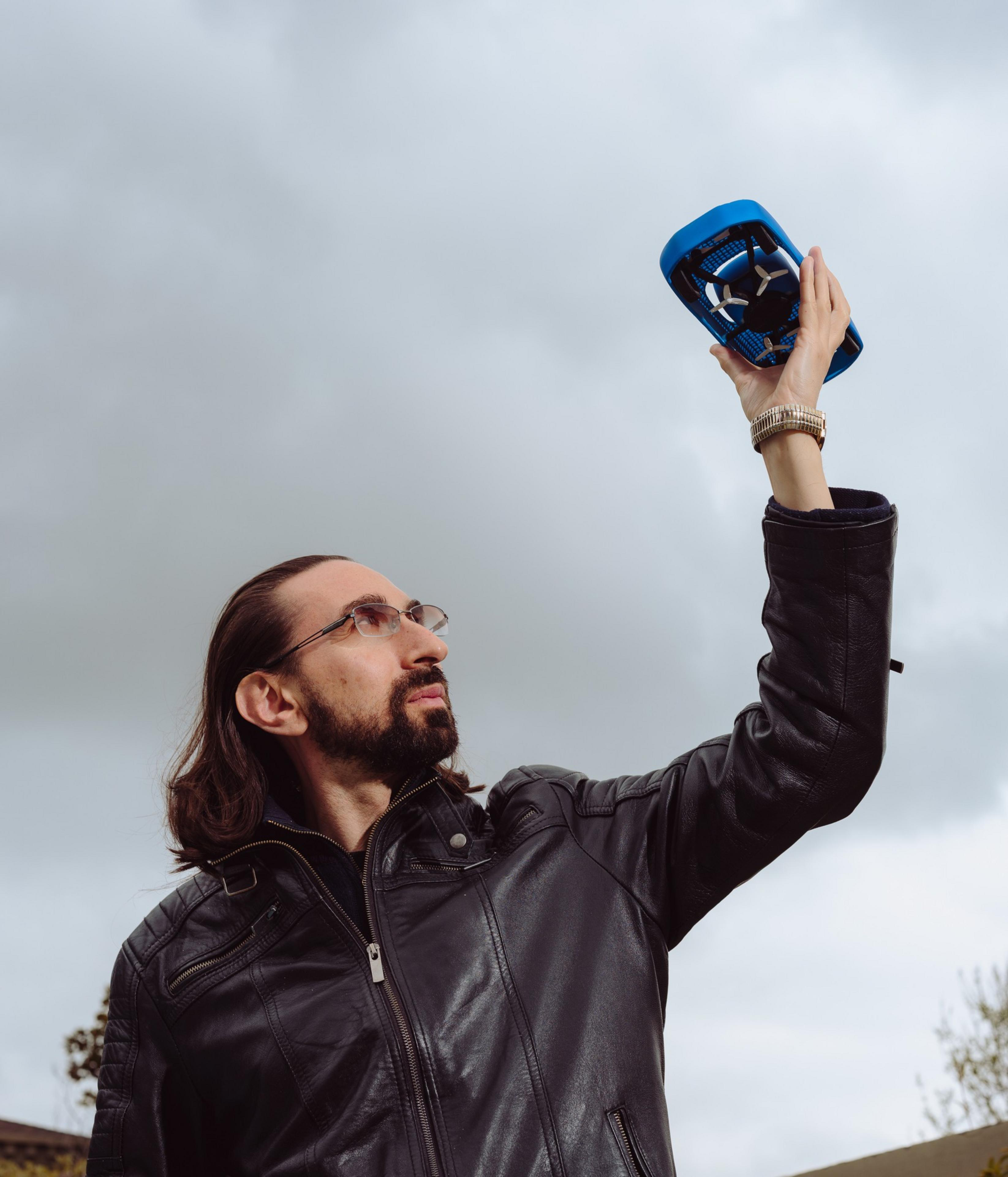 A person with long hair and glasses, wearing a leather jacket, is holding a small blue drone up to the cloudy sky.