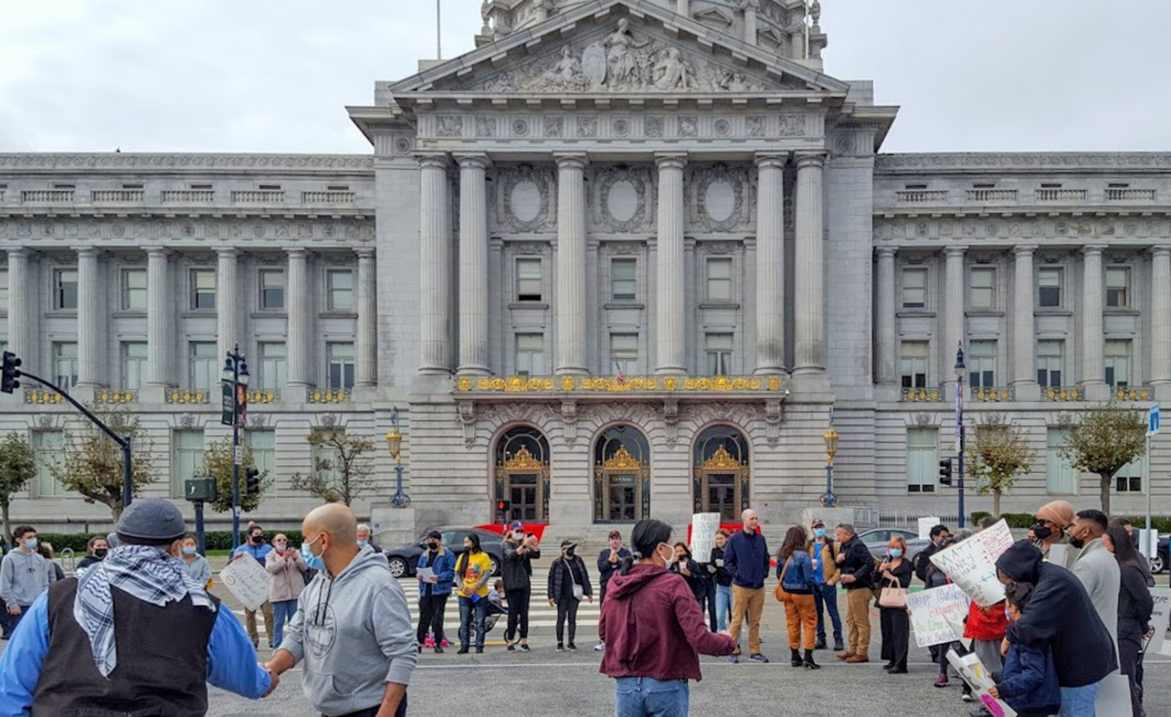 A group of people wearing masks gather in front of a large classical building with tall columns and ornate golden detailing for a protest or event.