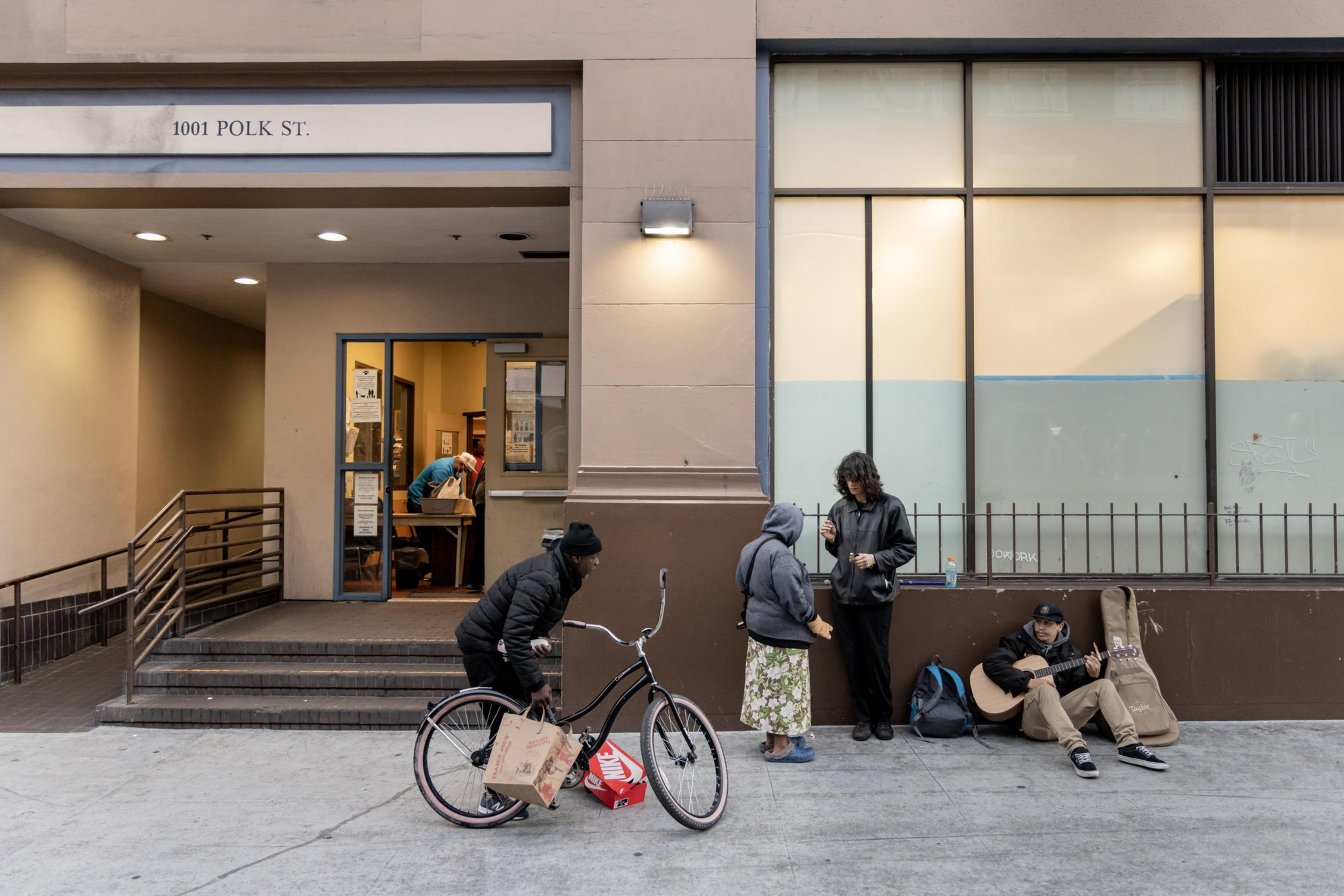 People are gathered outside a building at 1001 Polk St. One rides a bike holding a bag, two are talking, and another sits playing guitar. A person is seen inside.