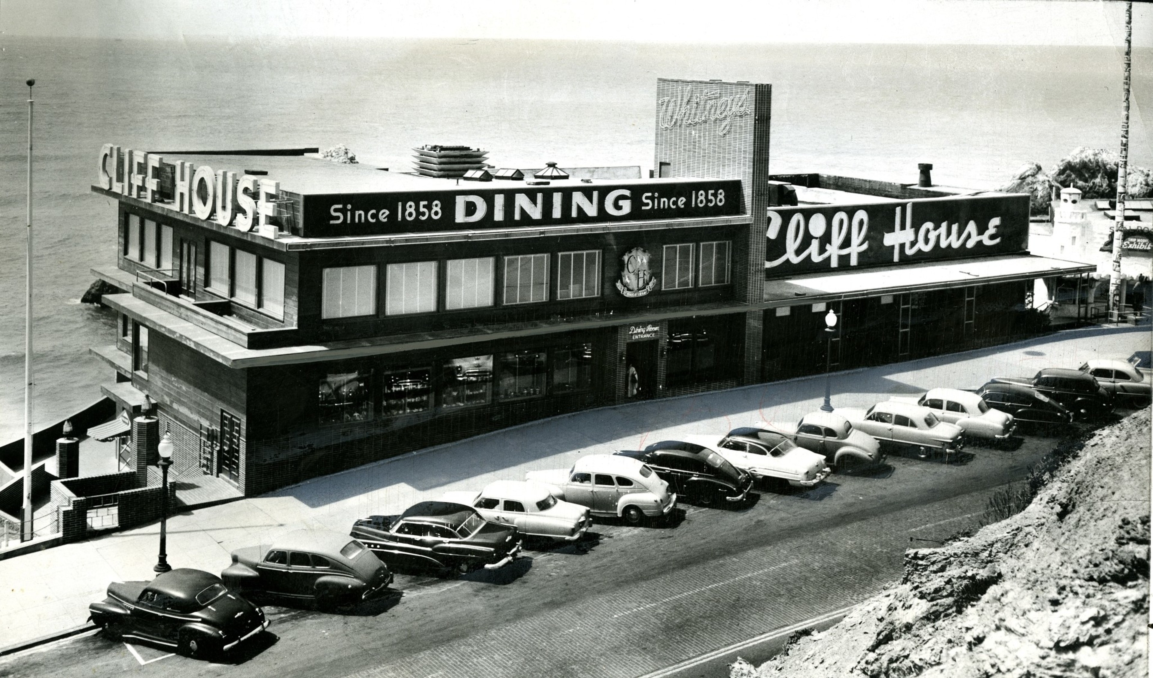 The black-and-white photo shows the Cliff House restaurant with vintage cars parked outside along a coastal road. The sign reads “Since 1858 DINING.”