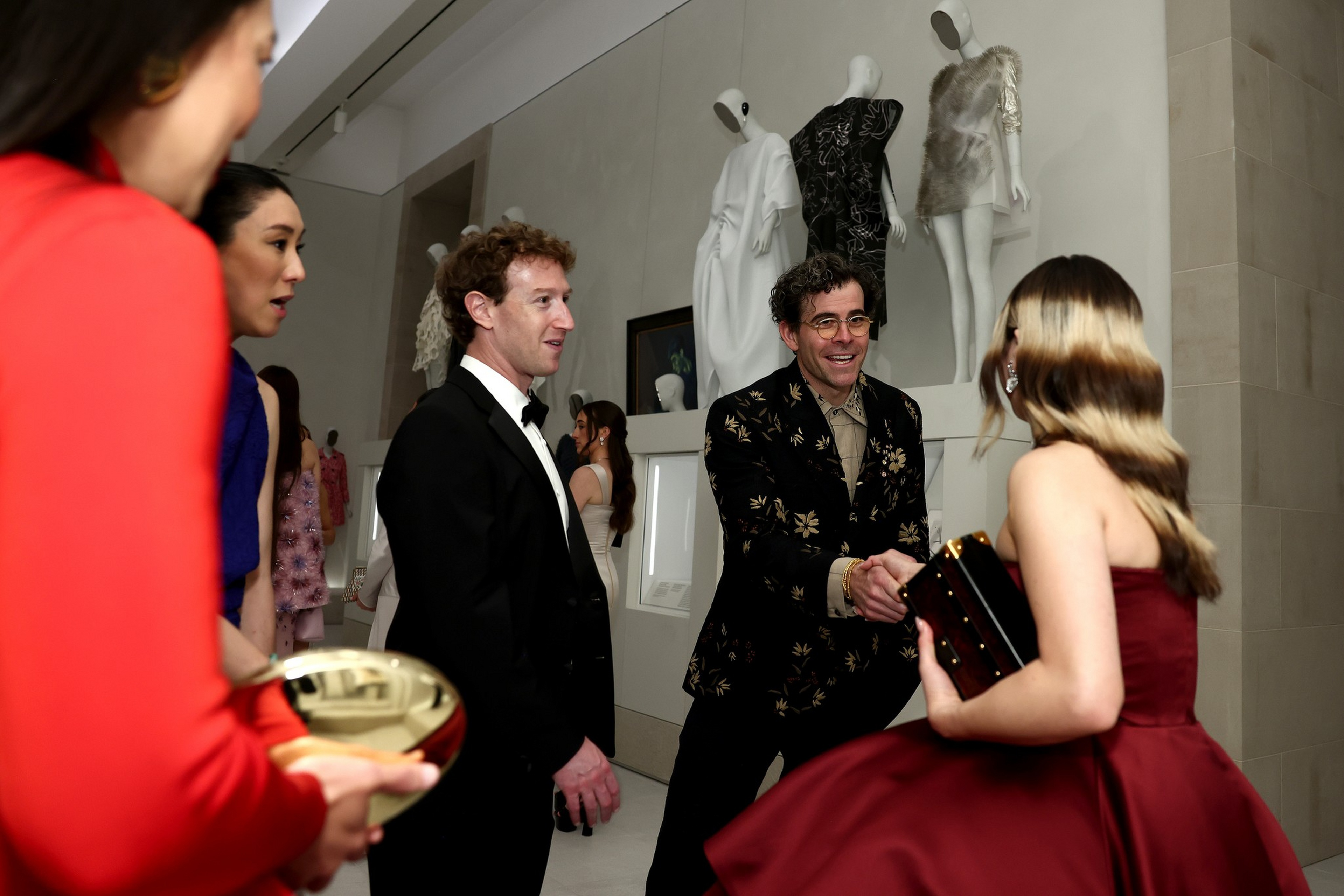 A man in a patterned black suit warmly shakes hands with a woman in a red dress while others look on in an indoor setting with white mannequins in the background.