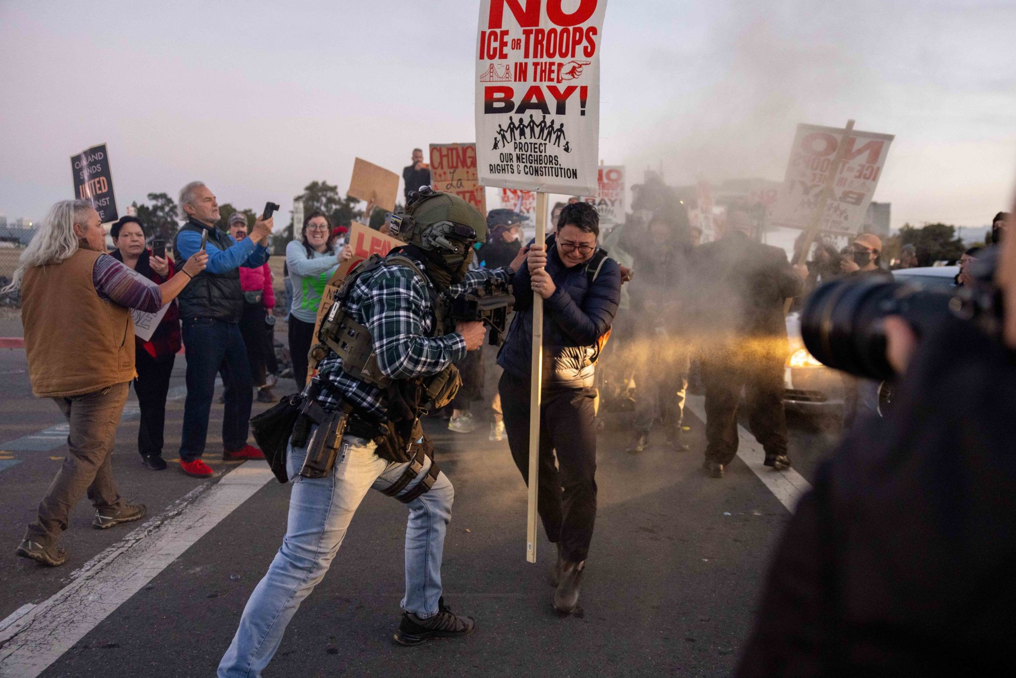 A protester holding a sign is sprayed with pepper spray by a heavily armed officer, while other protesters watch and capture the moment on their phones.