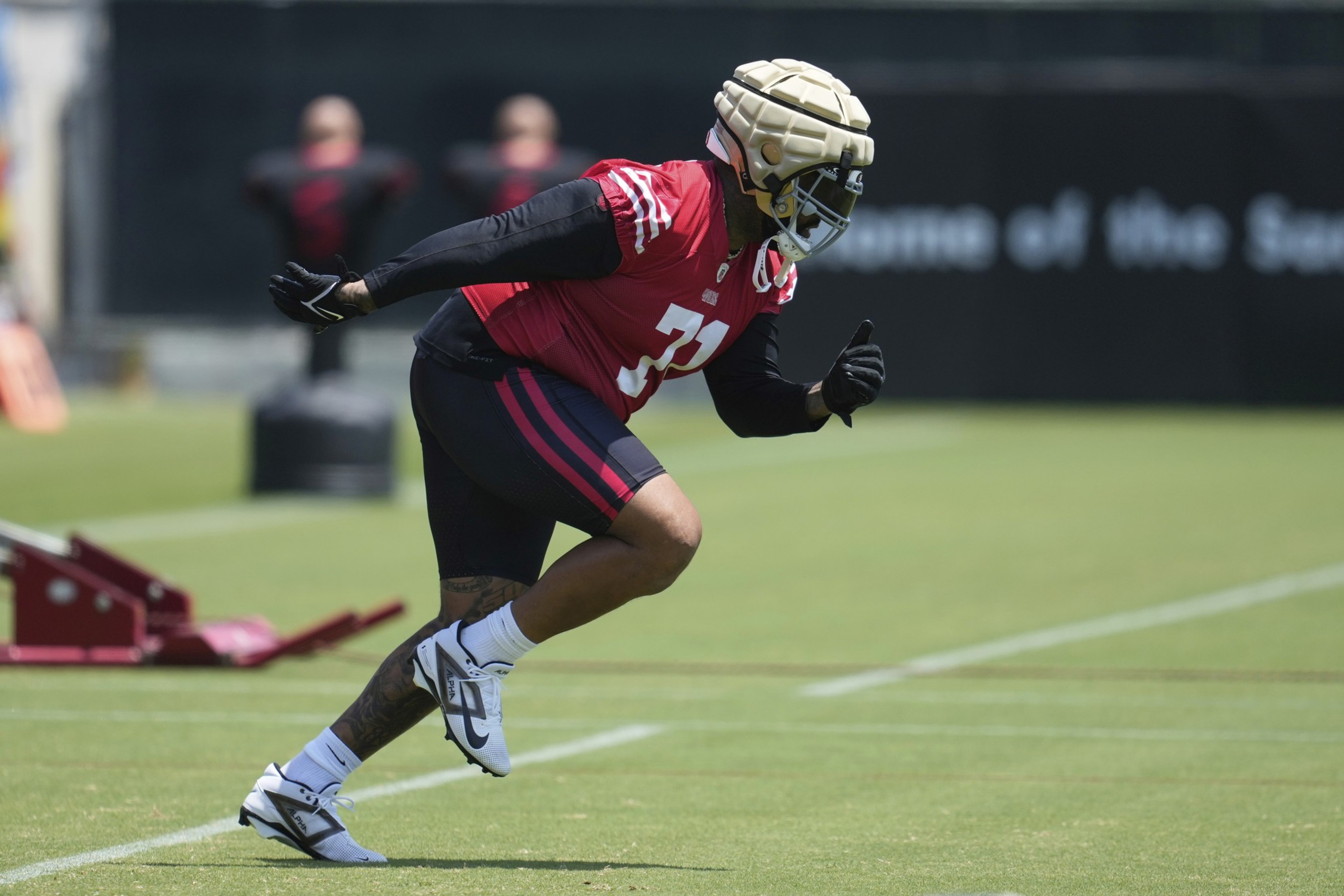 A football player wearing a padded helmet and red jersey runs on a grassy field during practice, with blurred training equipment in the background.