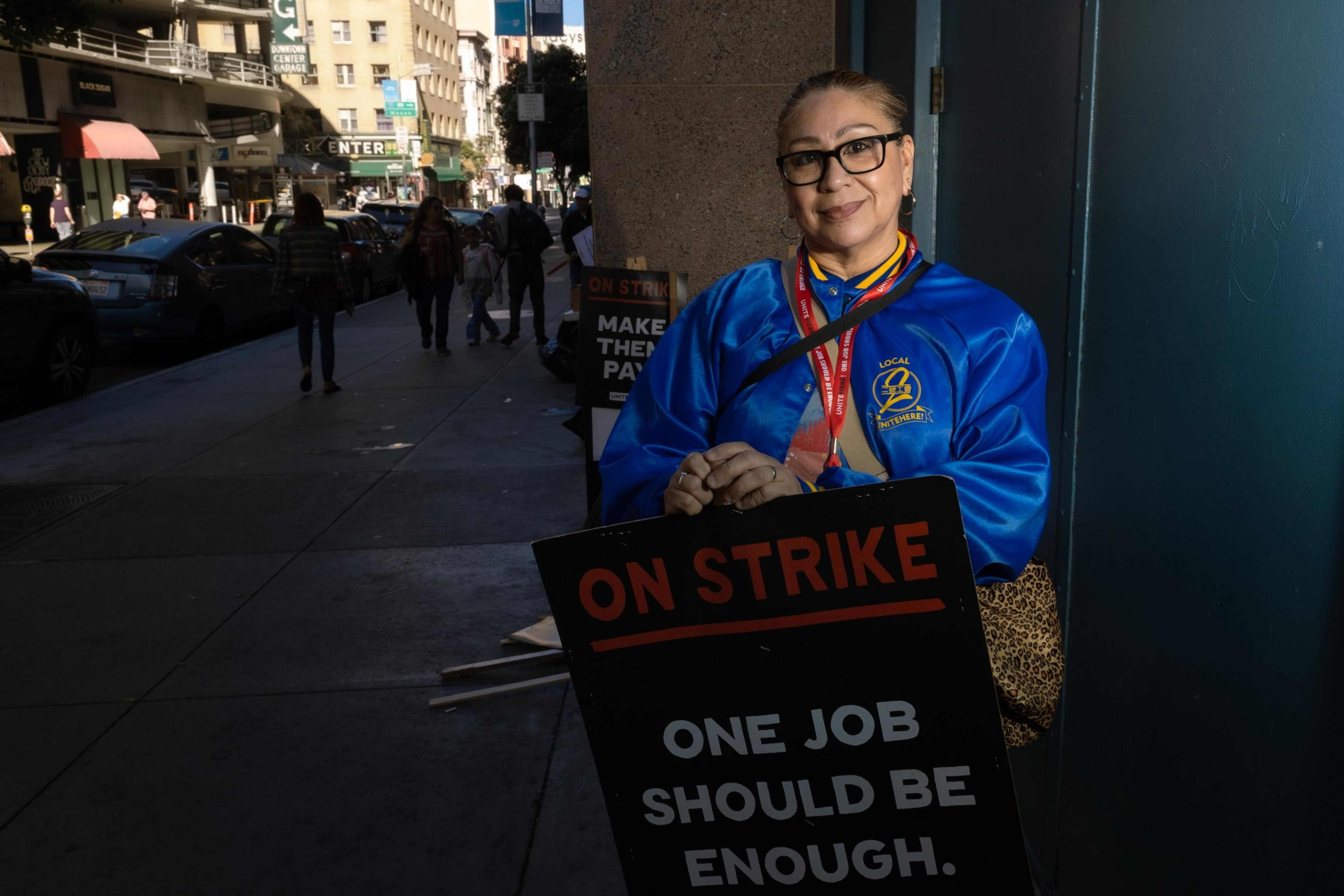 A woman in glasses and a blue jacket holds an &quot;ON STRIKE&quot; sign that reads &quot;One job should be enough.&quot; She's standing on a busy urban sidewalk.