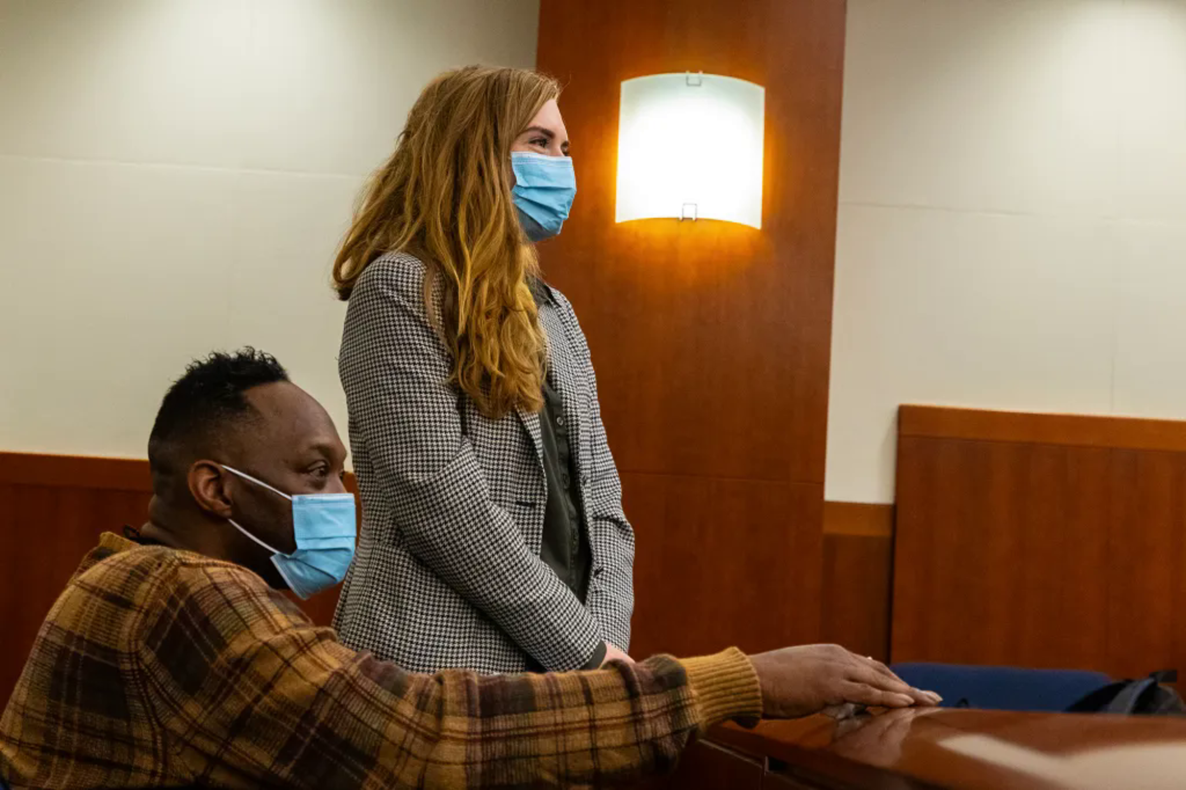 Woman wearing masks stands in courtroom next to man in mask sitting in pew