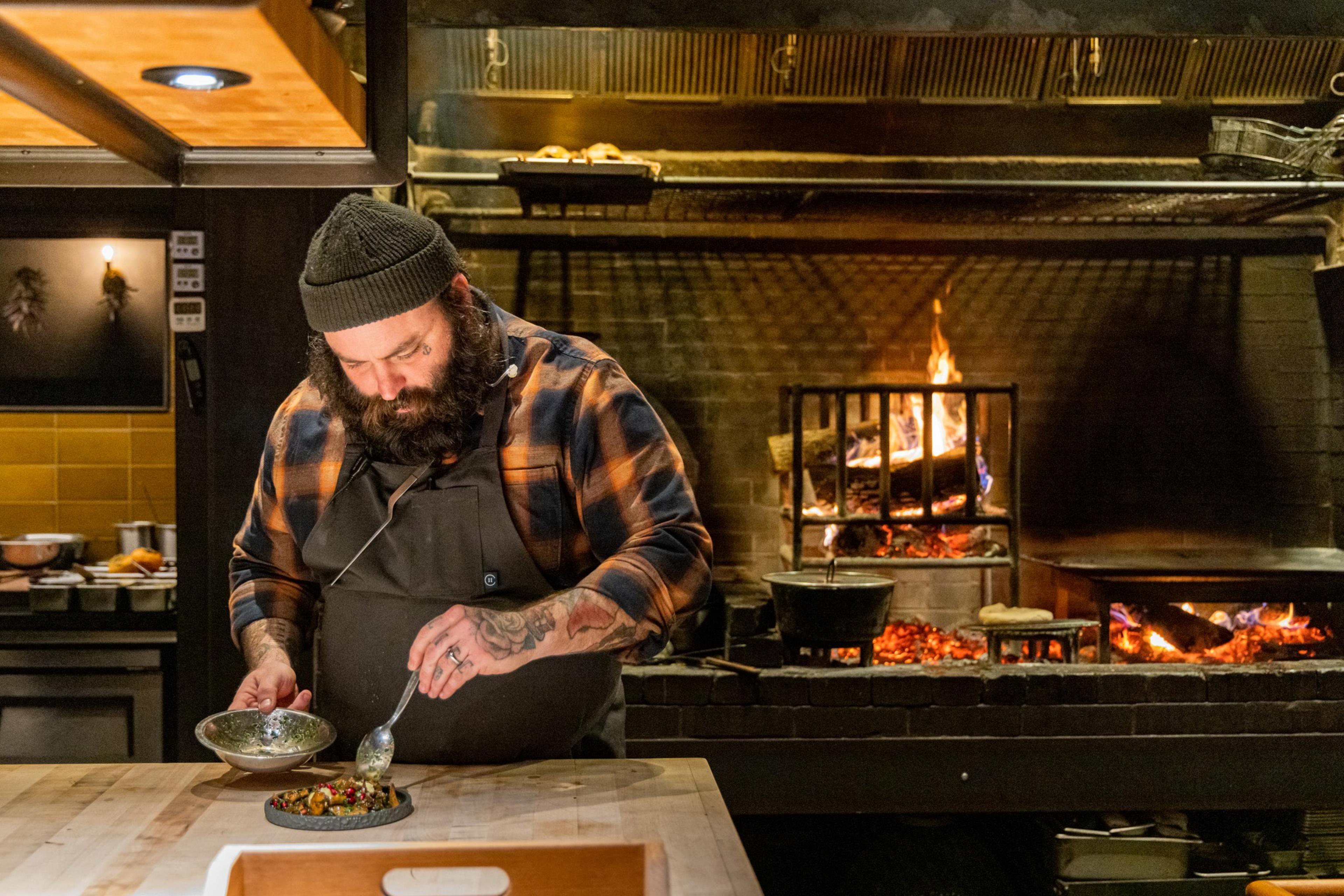 A bearded chef, wearing a beanie and apron, is plating food in a rustic kitchen with an open fire in the background.