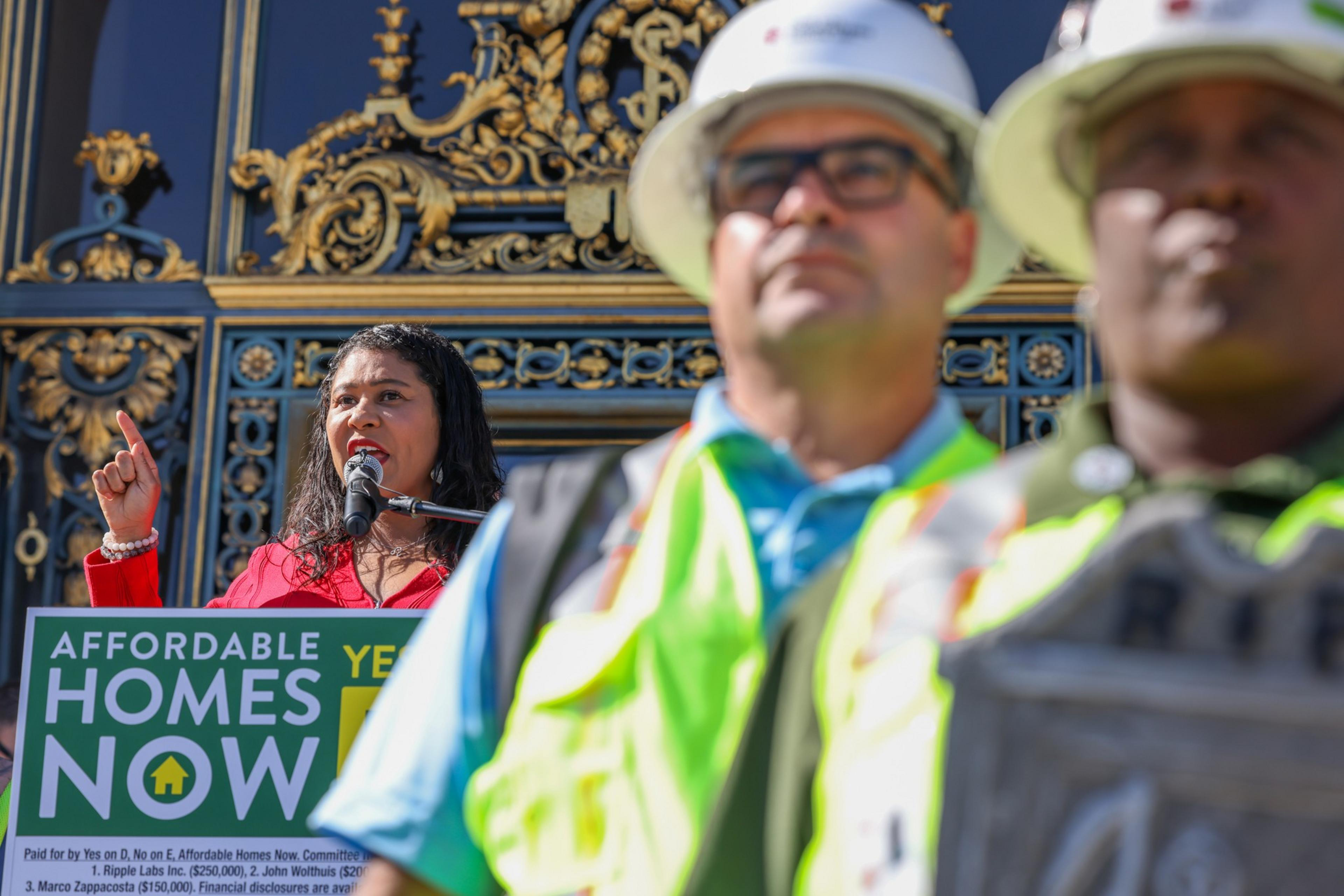 A woman speaks at a podium labeled "AFFORDABLE HOMES NOW YES" while two construction workers in hard hats and vests stand in the foreground. Ornate architecture is behind them.