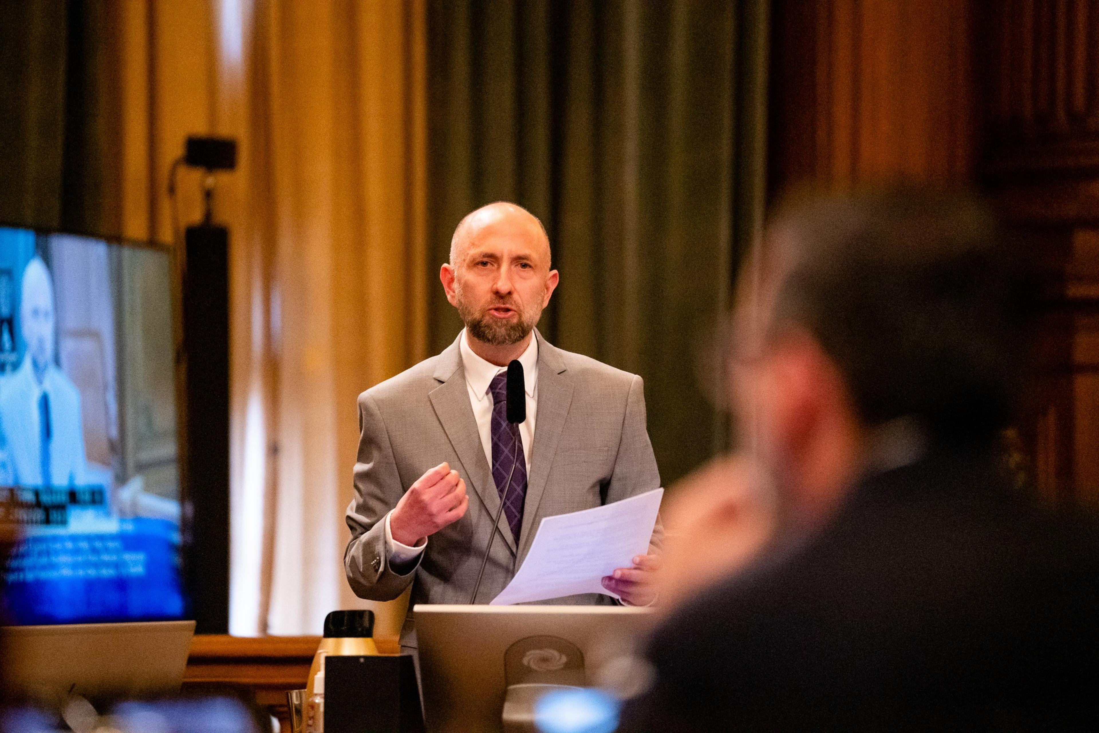 A man stands at a podium speaking into a microphone while gesticulating in the direction of an person.