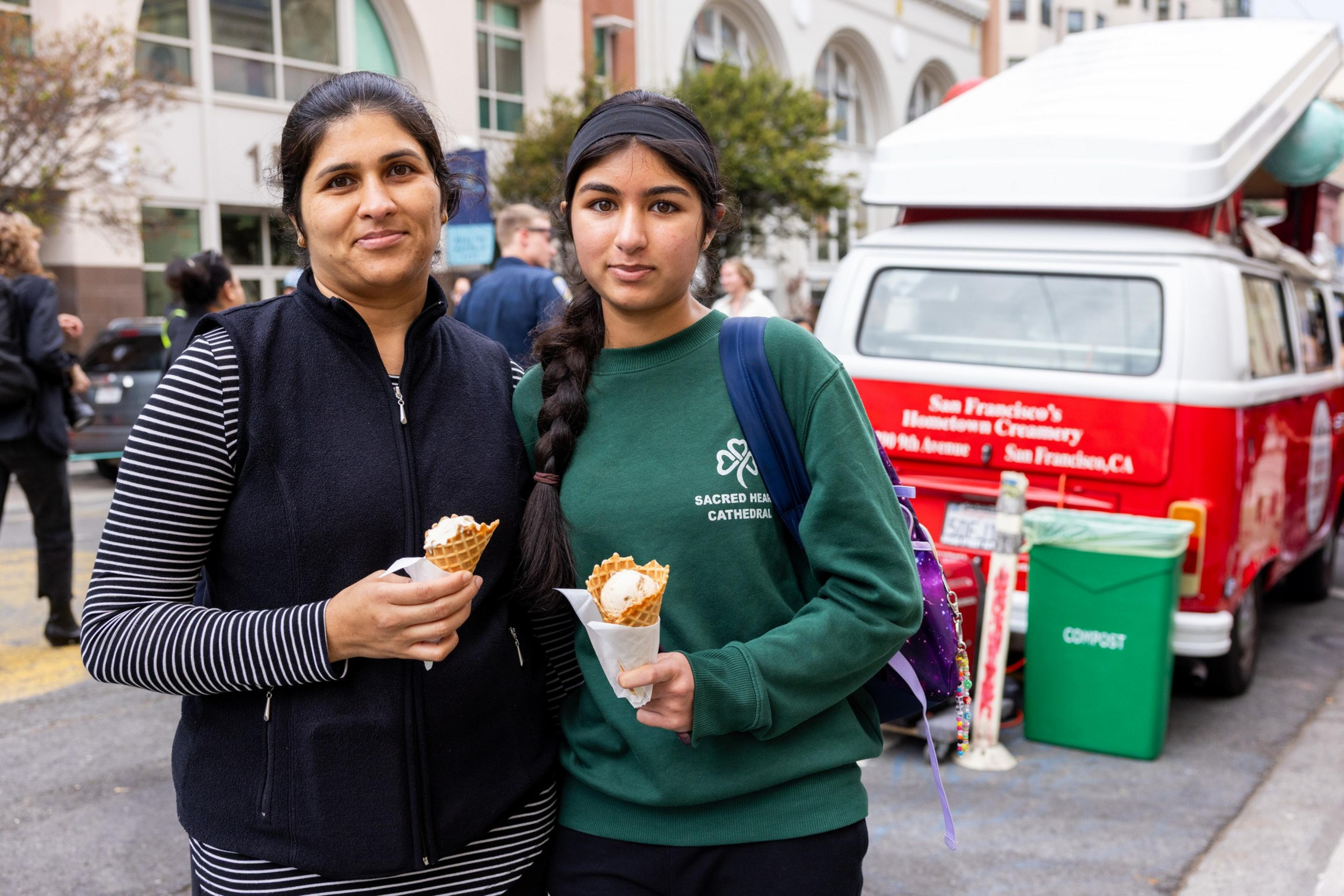 A woman and a girl stand side by side holding waffle cones with ice cream, with a red and white van and urban buildings in the background.