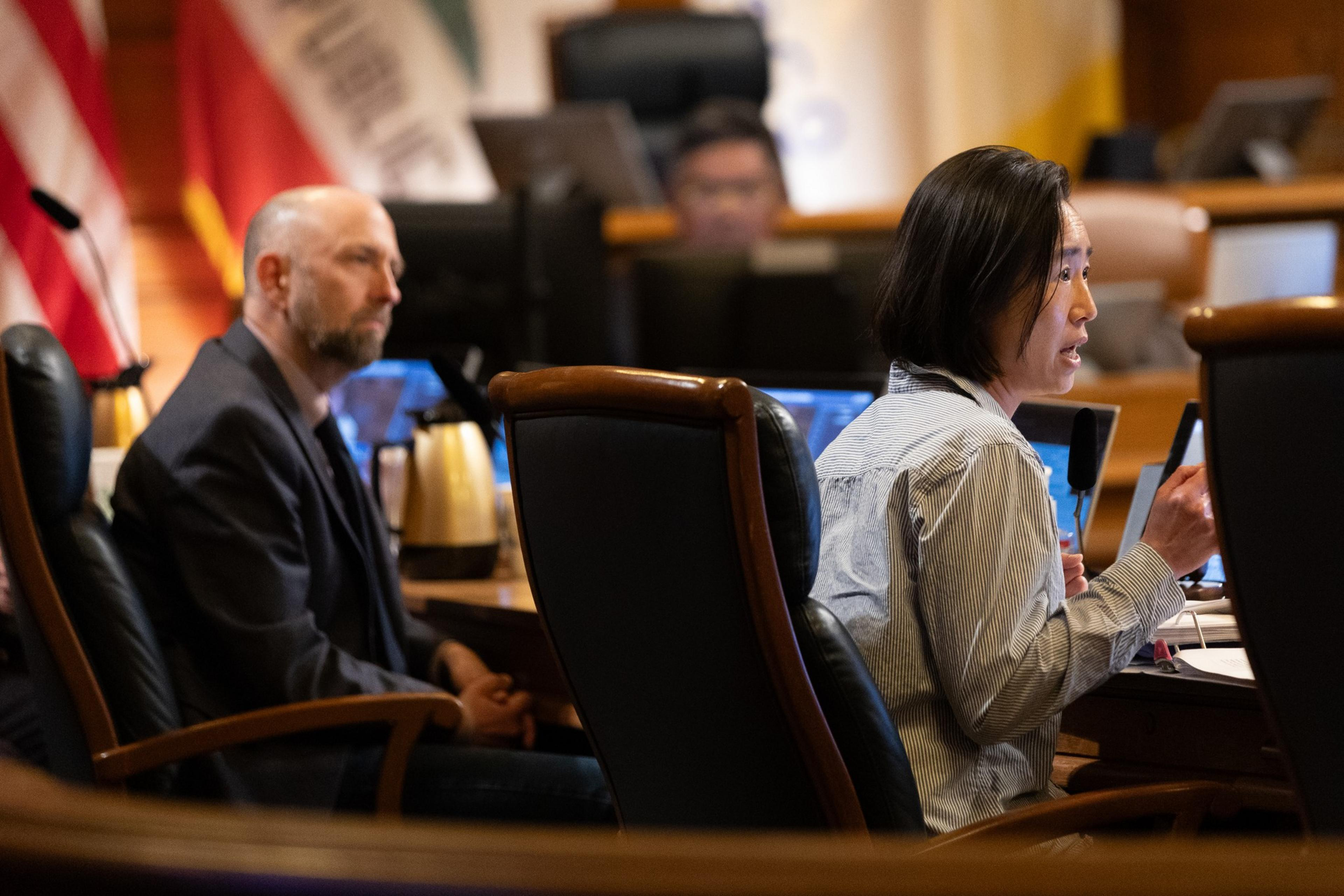 The image shows two people seated at a table in a formal setting. One person speaks into a microphone, while the other listens attentively. Flags are visible in the background.