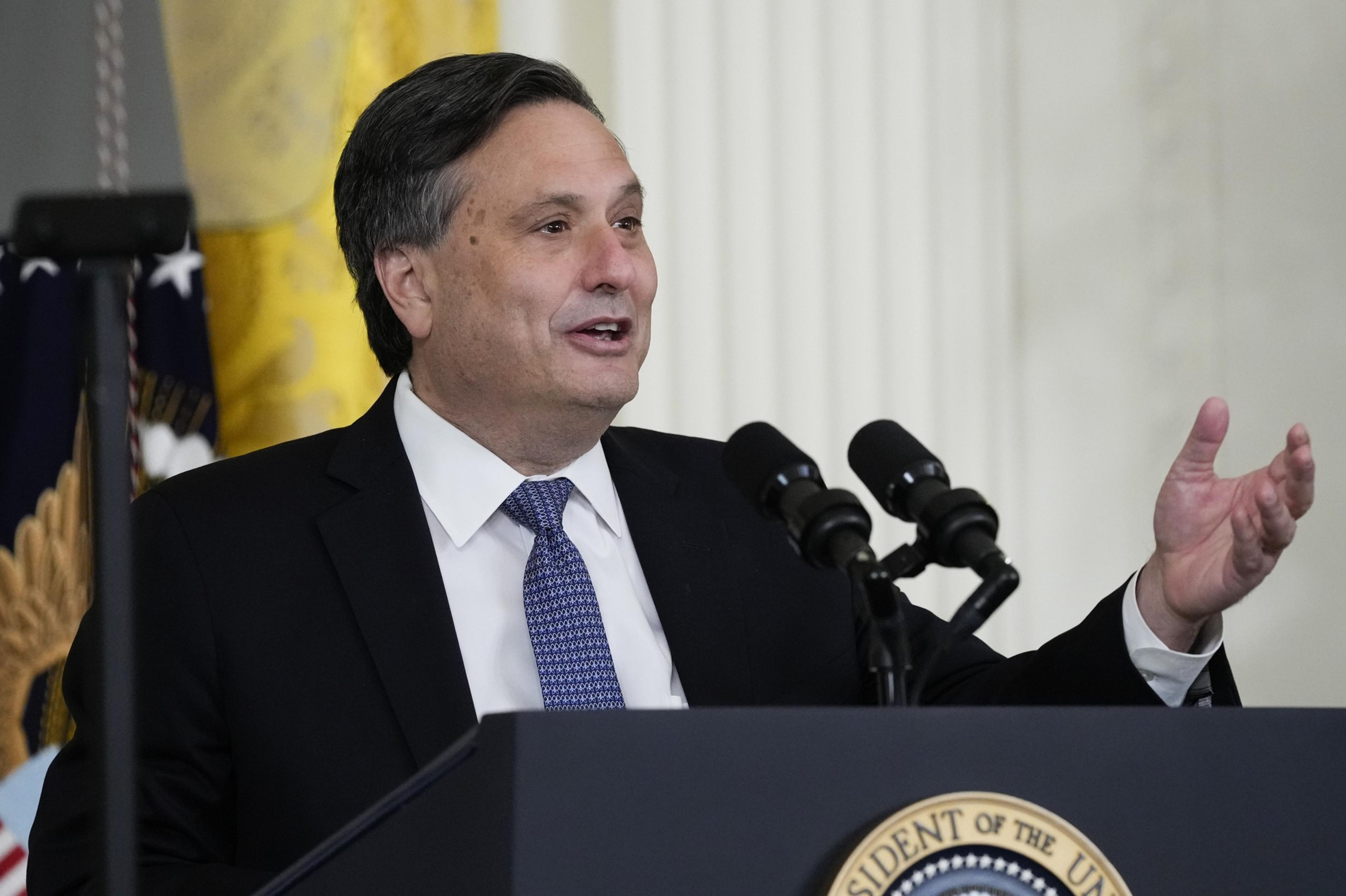 A suit-wearing man stands at a podium with a presidential seal in front of it and waves his left hand slightly.