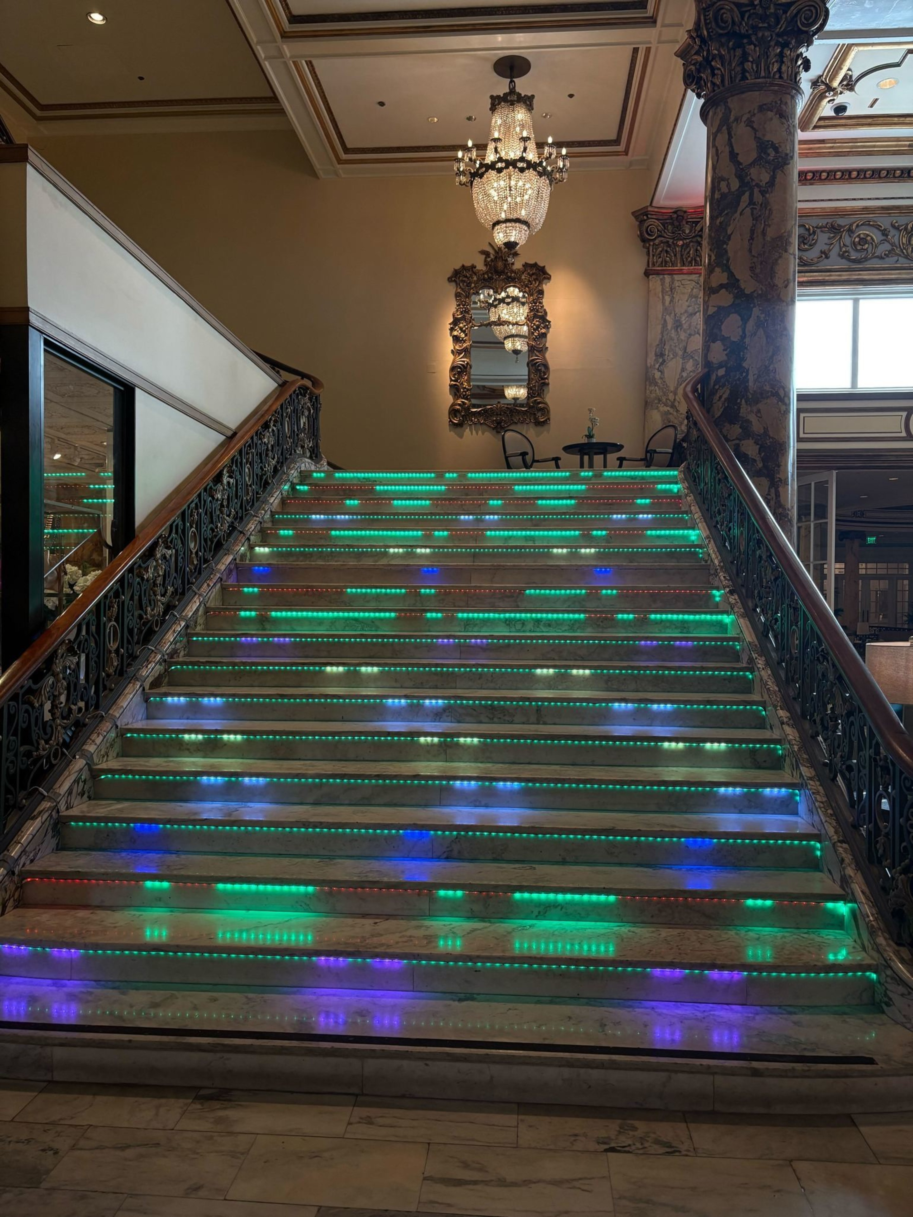A grand marble staircase with ornate iron railings features colorful LED lights on each step, leading up to a landing with a chandelier and ornate mirror.