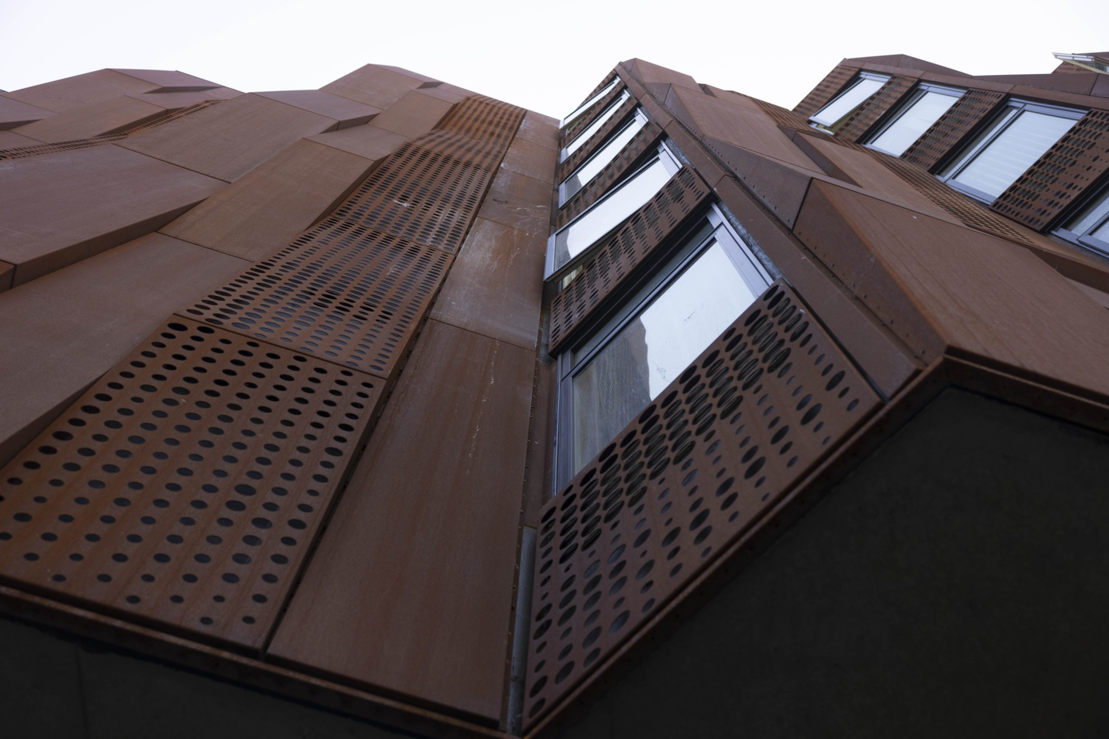 The image shows an upward view of a modern building facade with rust-colored panels and large windows, featuring a grid of circular perforations.