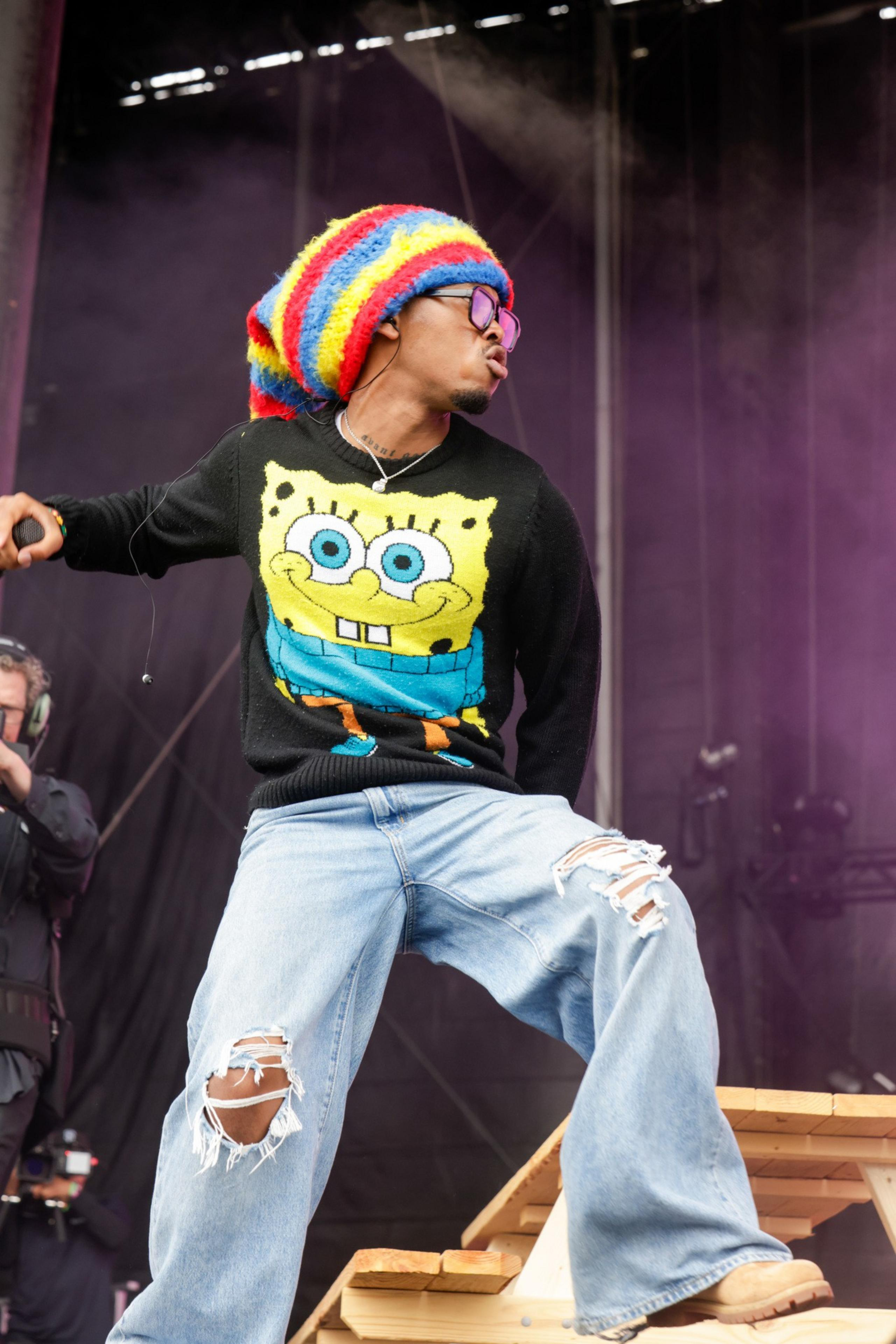 A person wearing a rainbow hat, SpongeBob sweatshirt, ripped jeans, and sunglasses holds a microphone and performs energetically on stage.