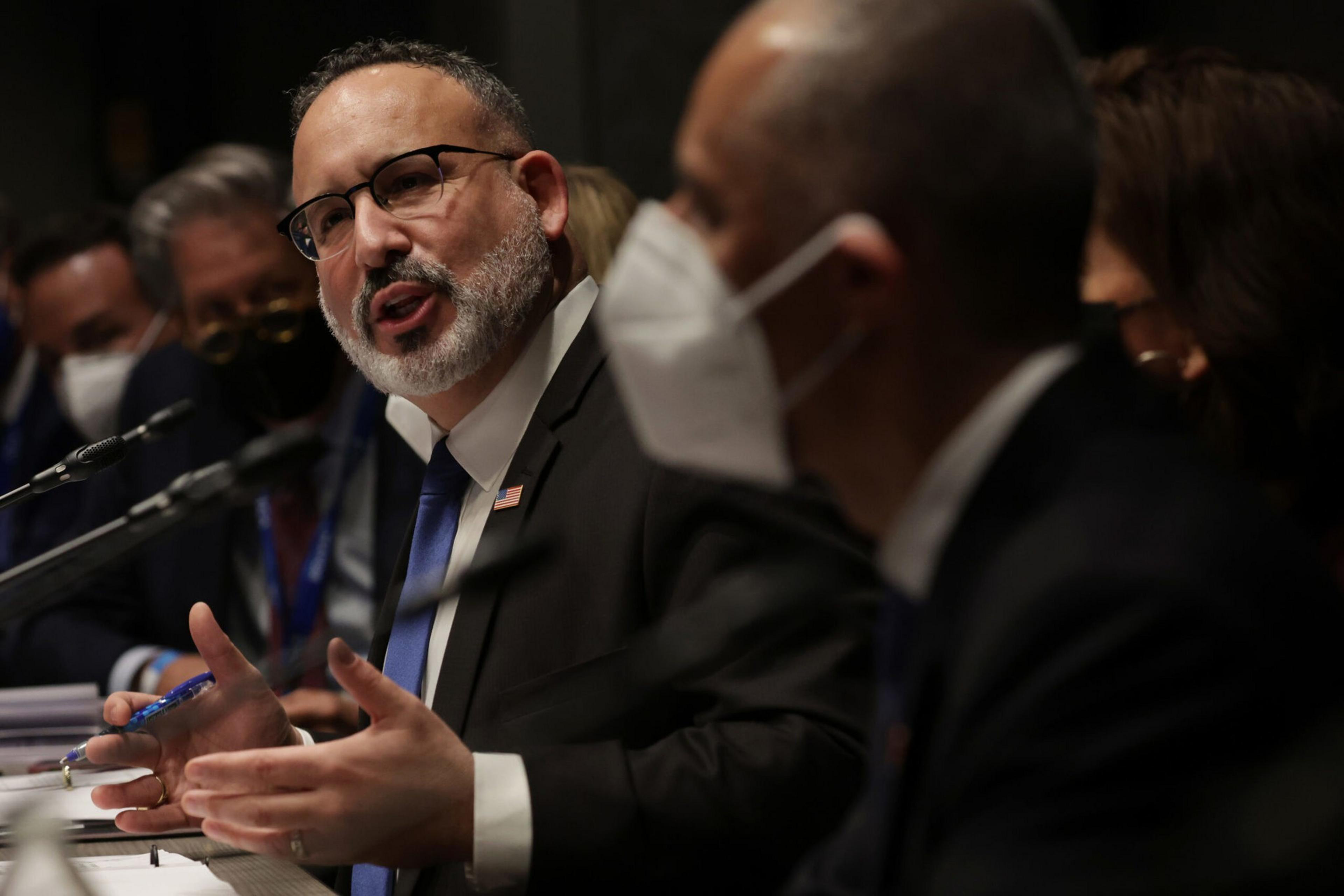 A man with glasses and a beard speaks while gesturing with his hands during a formal meeting, with others beside him wearing masks.