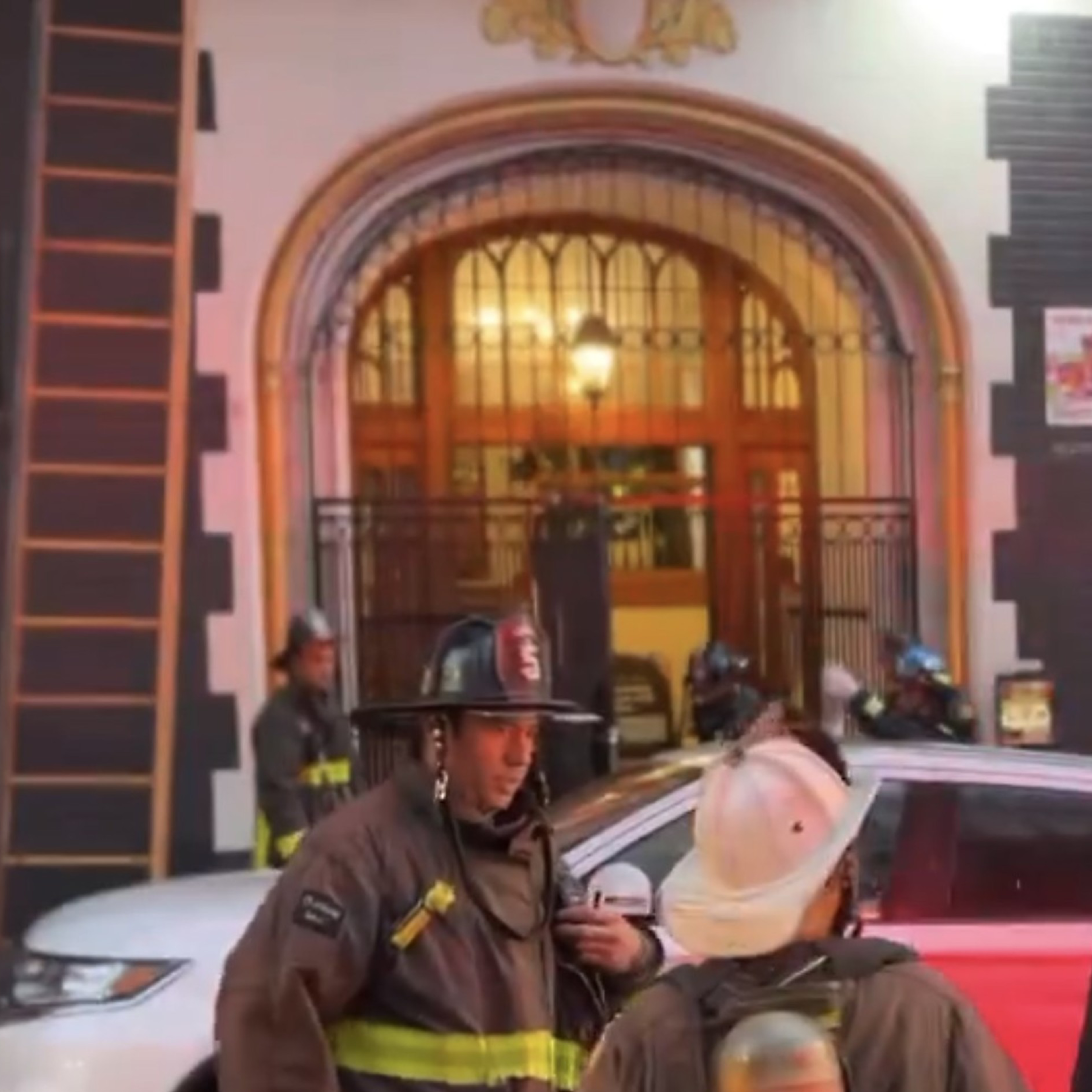 Three firefighters in gear stand near vehicles on a street in front of a building with an arched entrance. Other firefighters and equipment are visible in the background.