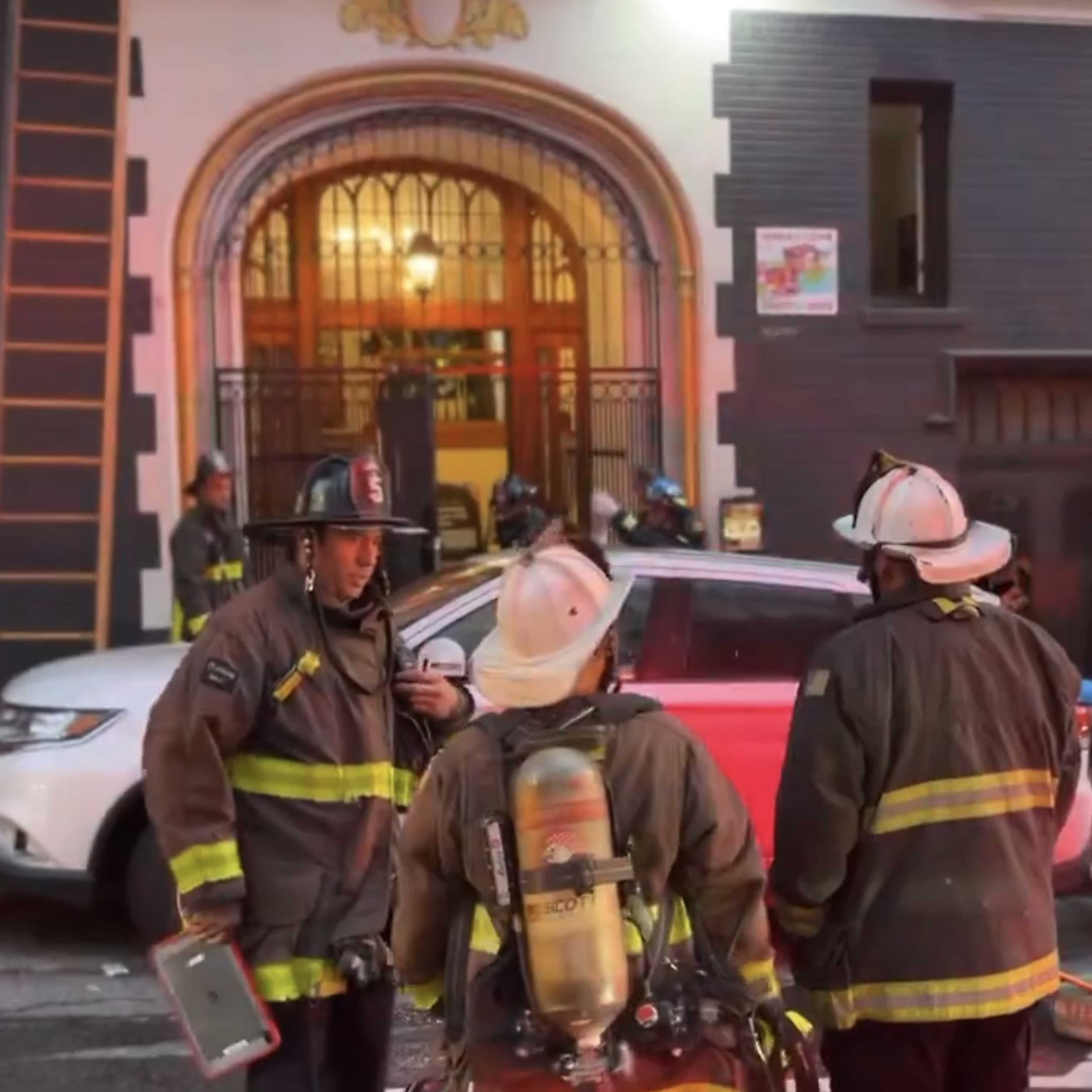 Three firefighters in gear stand near vehicles on a street in front of a building with an arched entrance. Other firefighters and equipment are visible in the background.