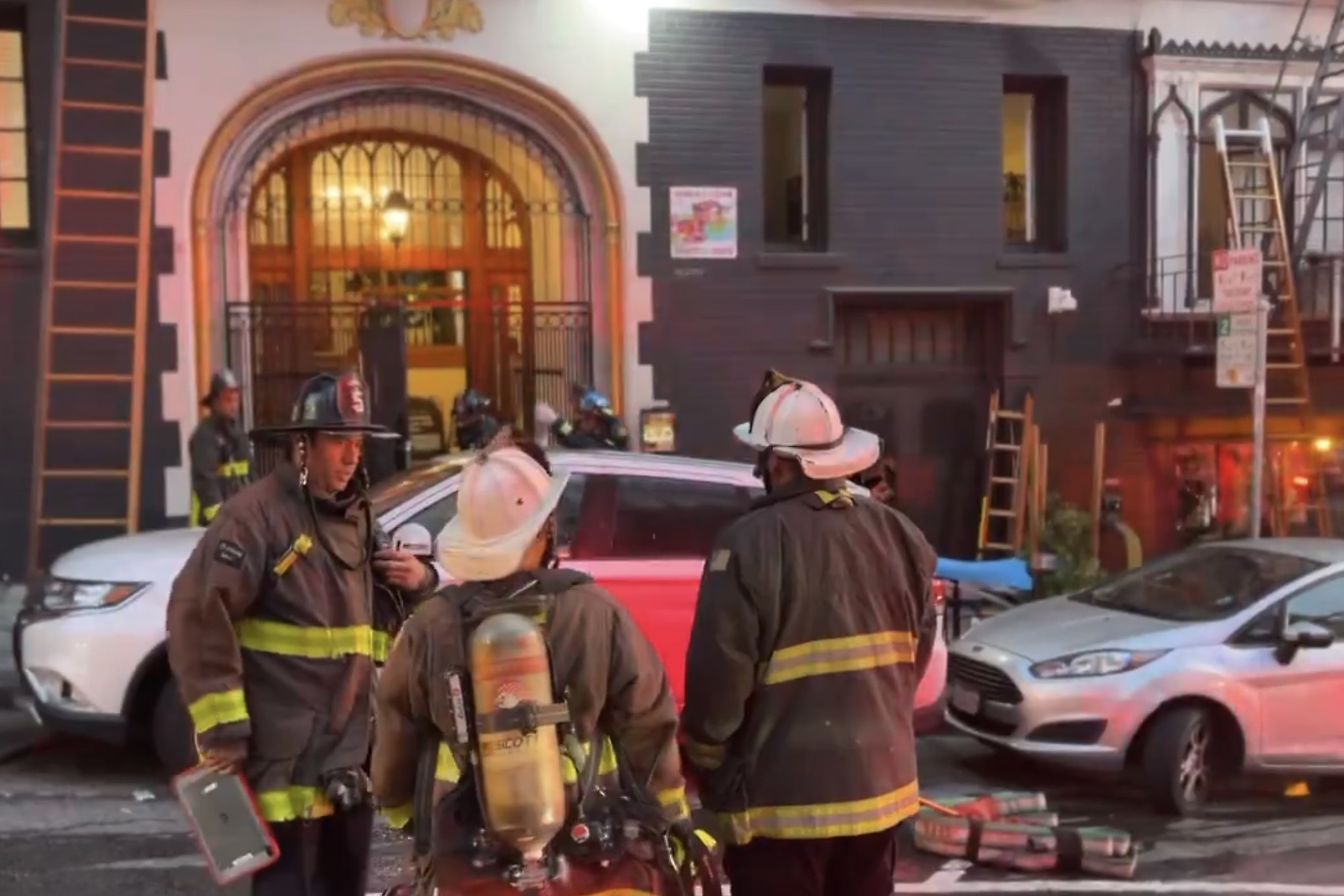 Three firefighters in gear stand near vehicles on a street in front of a building with an arched entrance. Other firefighters and equipment are visible in the background.