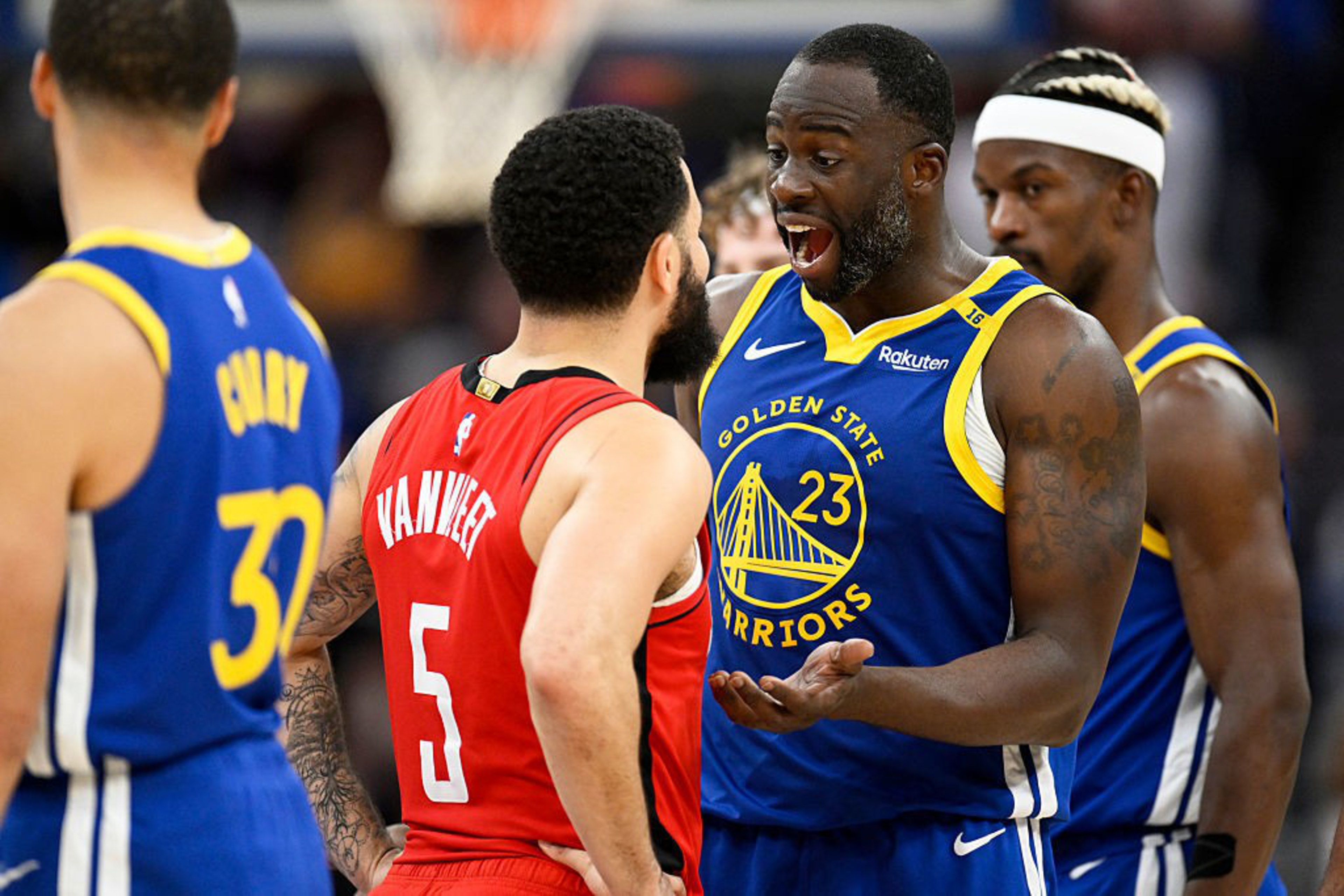 A basketball player in a blue Golden State Warriors jersey is animatedly talking to an opponent in a red jersey, with two other players nearby observing.