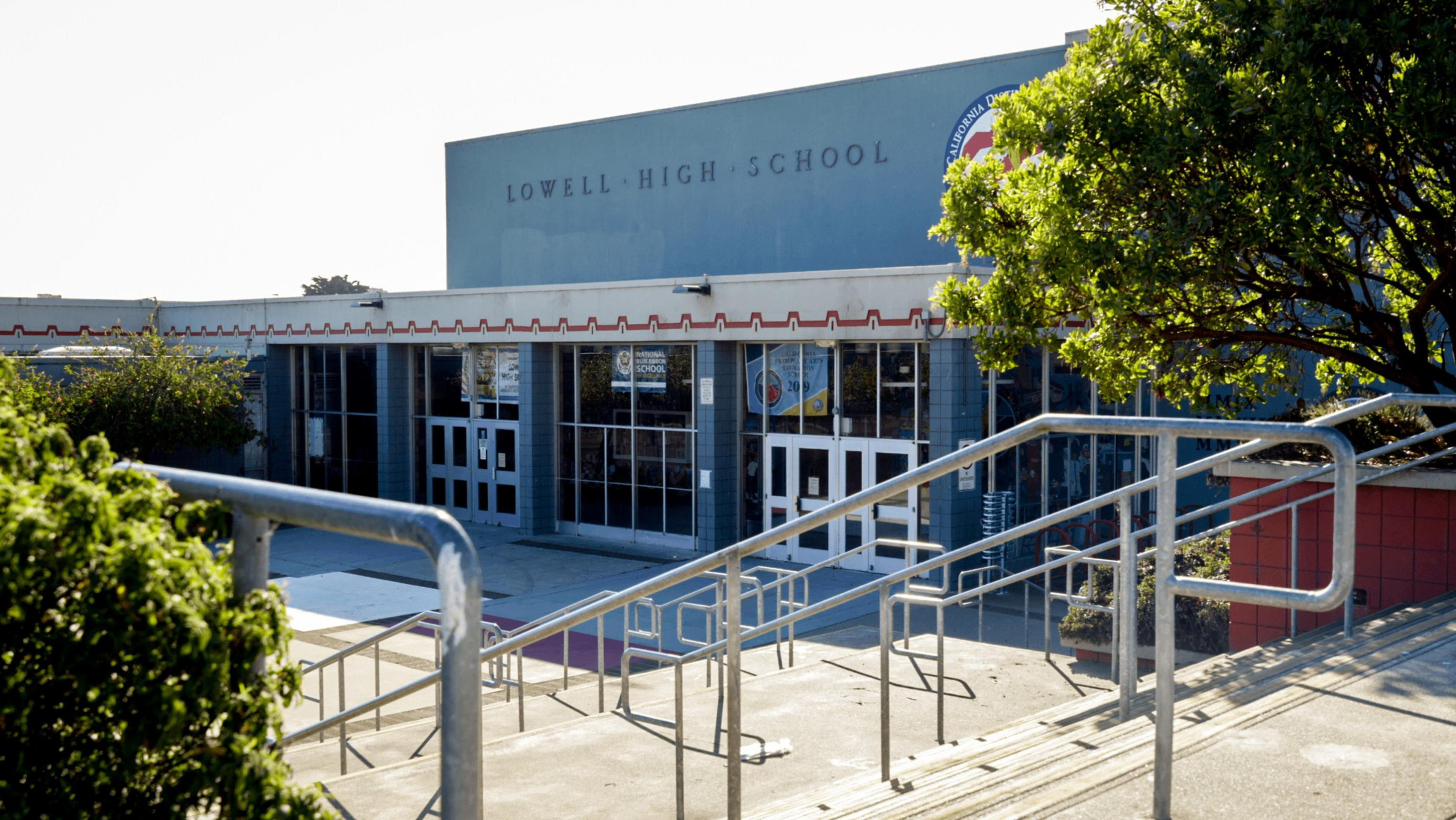 Lowell High School entrance with multiple doors and glass panels, surrounded by metal railings, stairs, bushes, and a tree on a sunny day.
