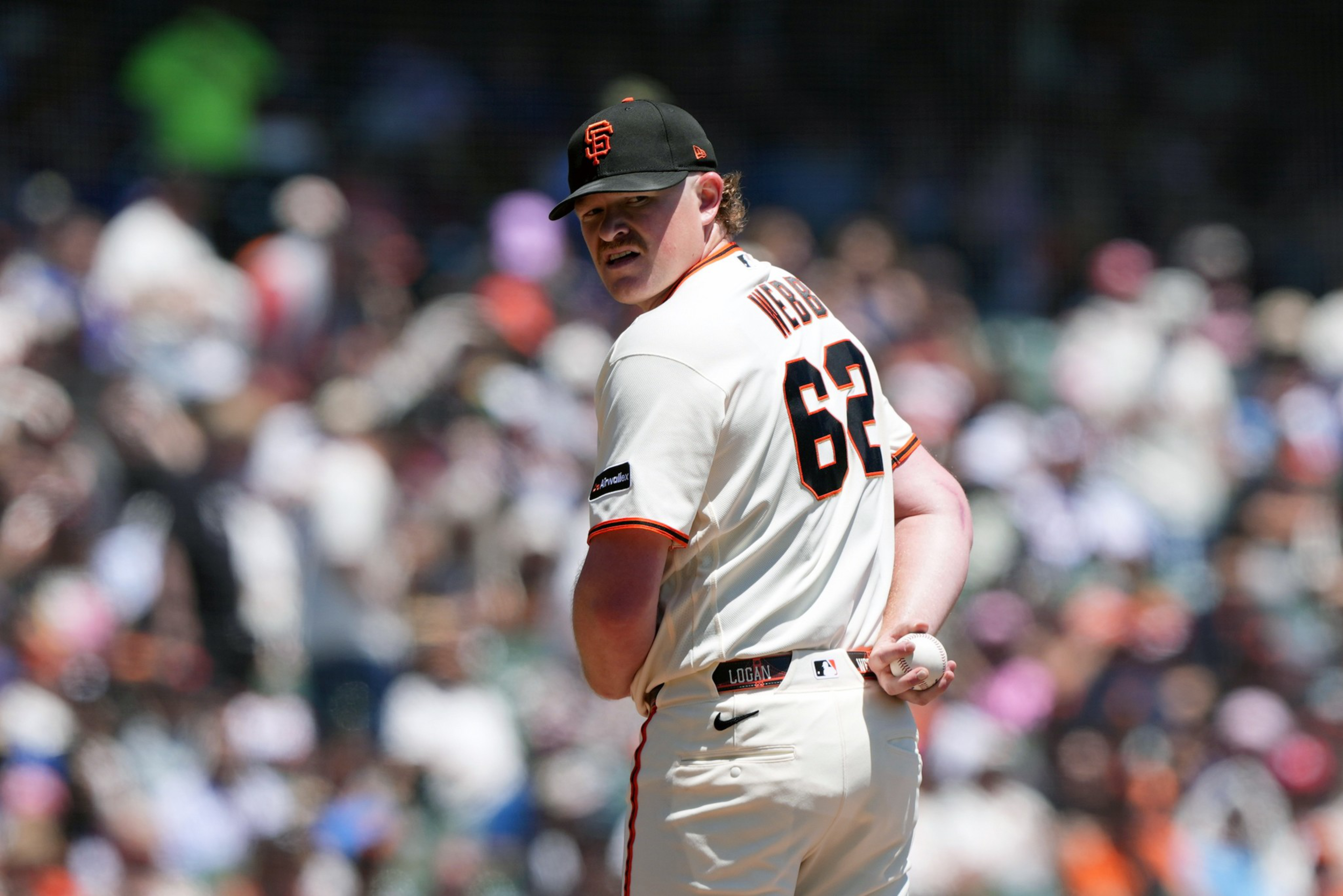 A baseball player wearing a white San Francisco Giants uniform with number 62 stands on the field holding a baseball behind his back.