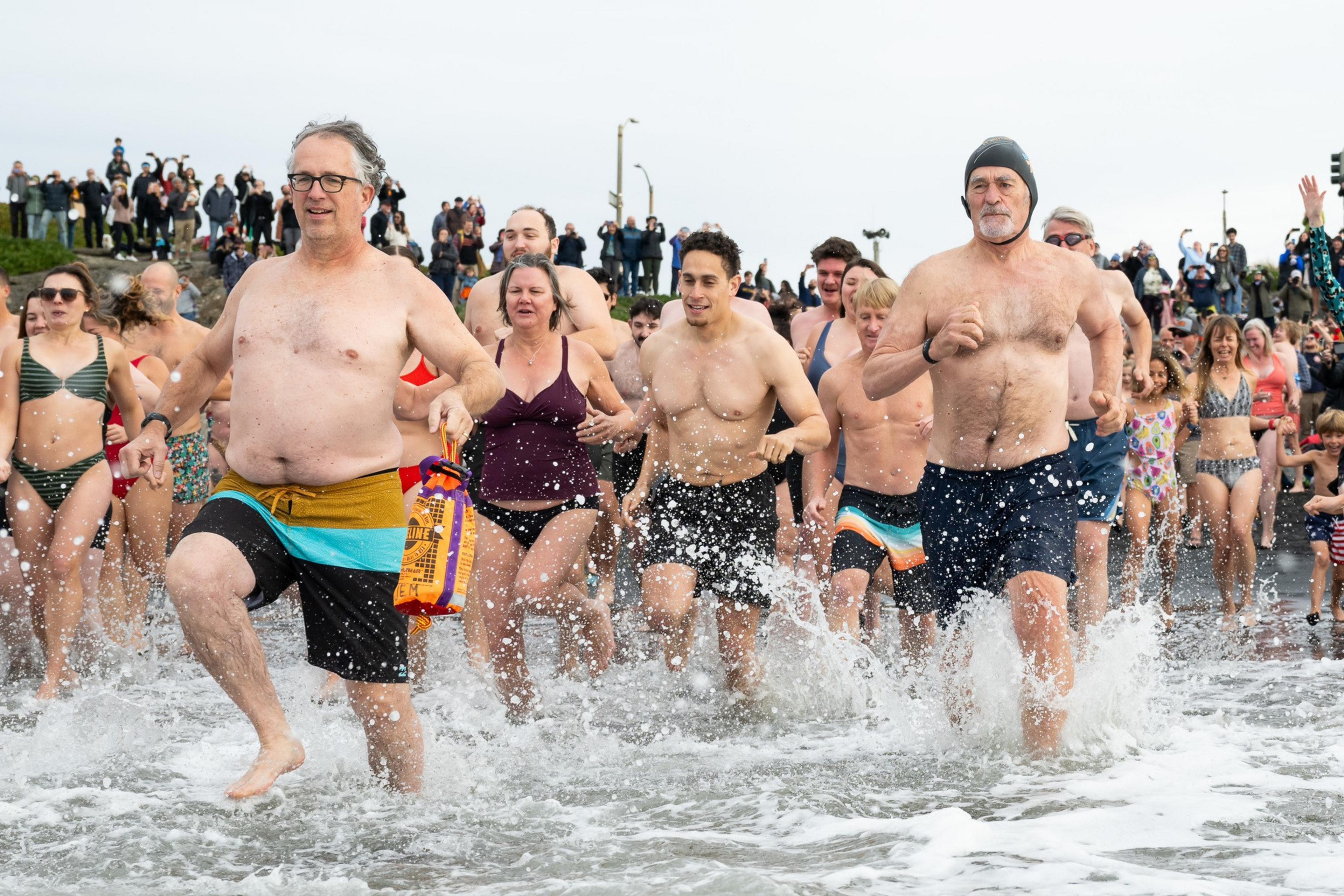 A large group of people in swimwear rushes into the sea, splashing water everywhere. Behind them, a crowd watches from the shore under an overcast sky.