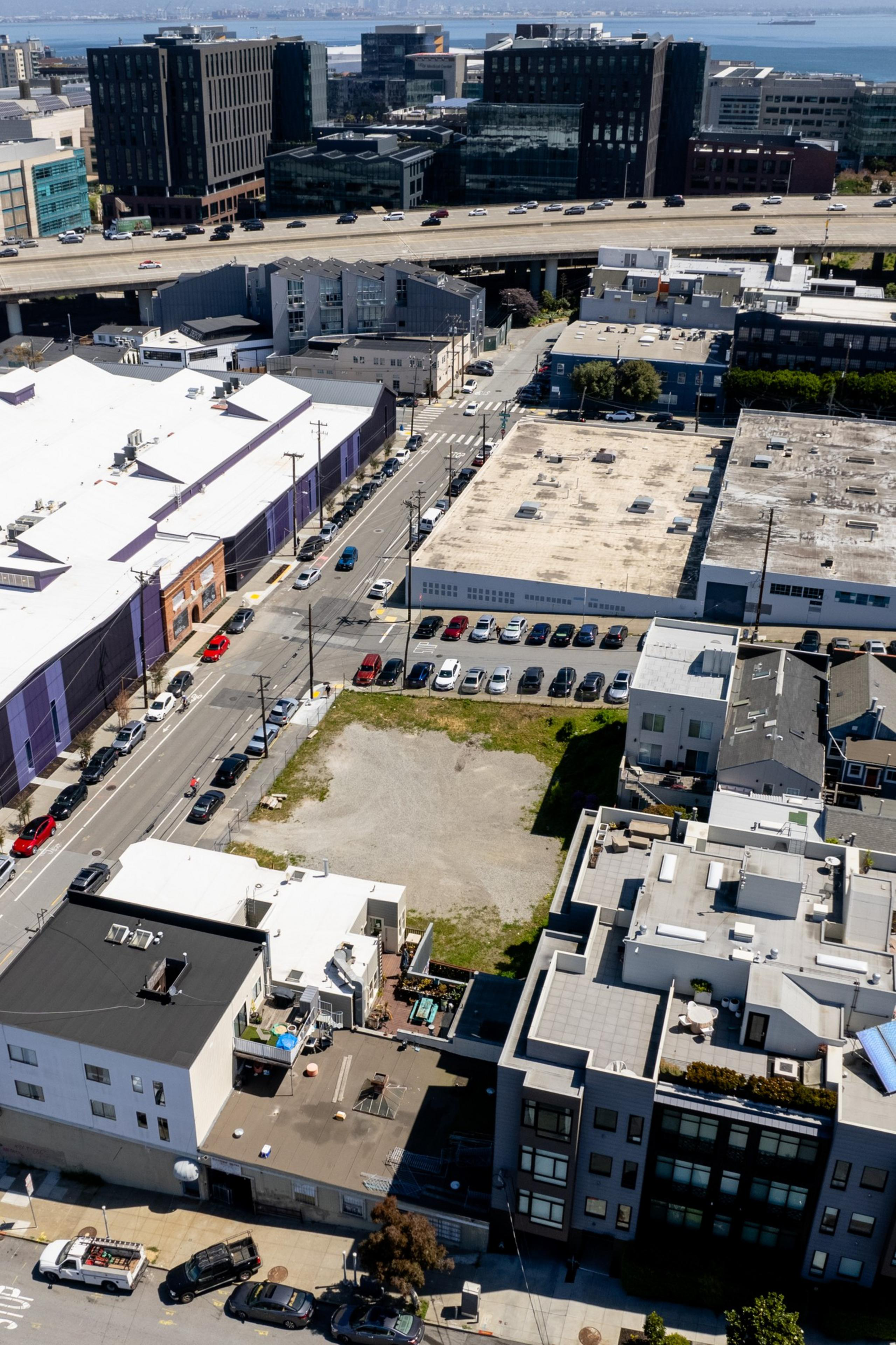 A bird's-eye view of an urban landscape with buildings, a highway in the distance, cars, and an open lot.