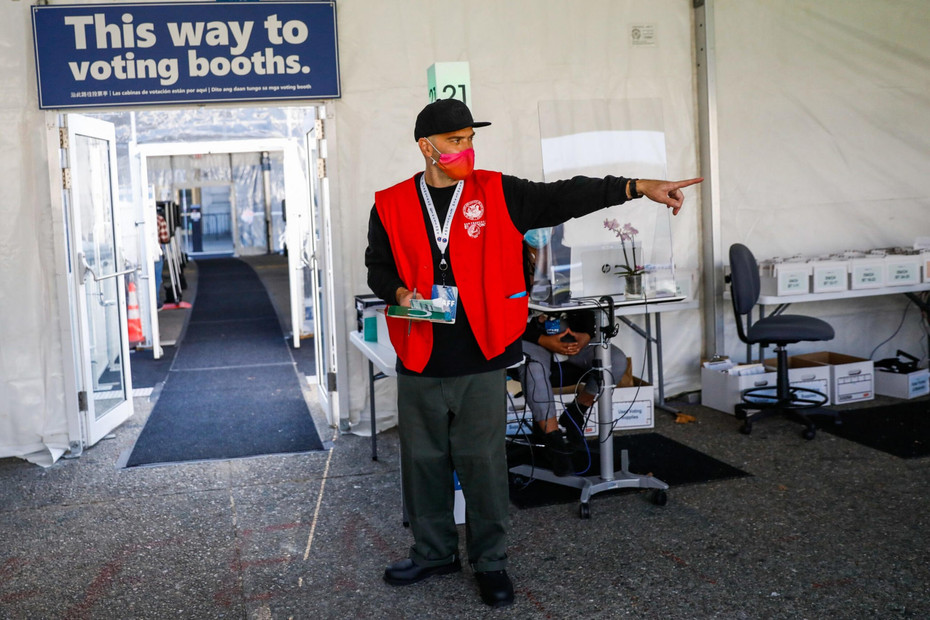 A person wearing a red vest and pink mask points to the right under a sign that says "This way to voting booths" at a voting site.