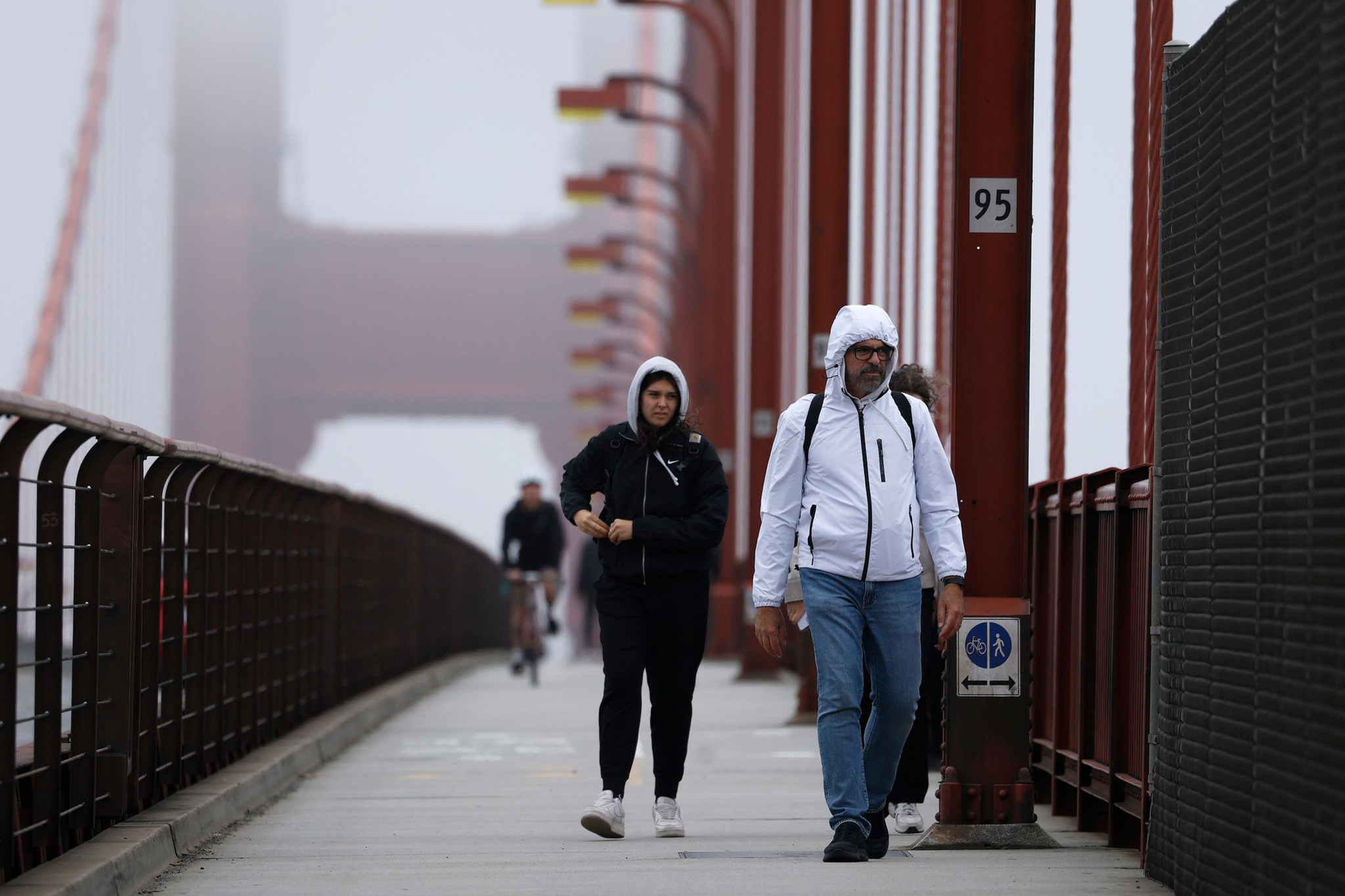 Two people in hooded jackets walk on a foggy Golden Gate Bridge sidewalk, with a cyclist in the background and red bridge structures on the right.