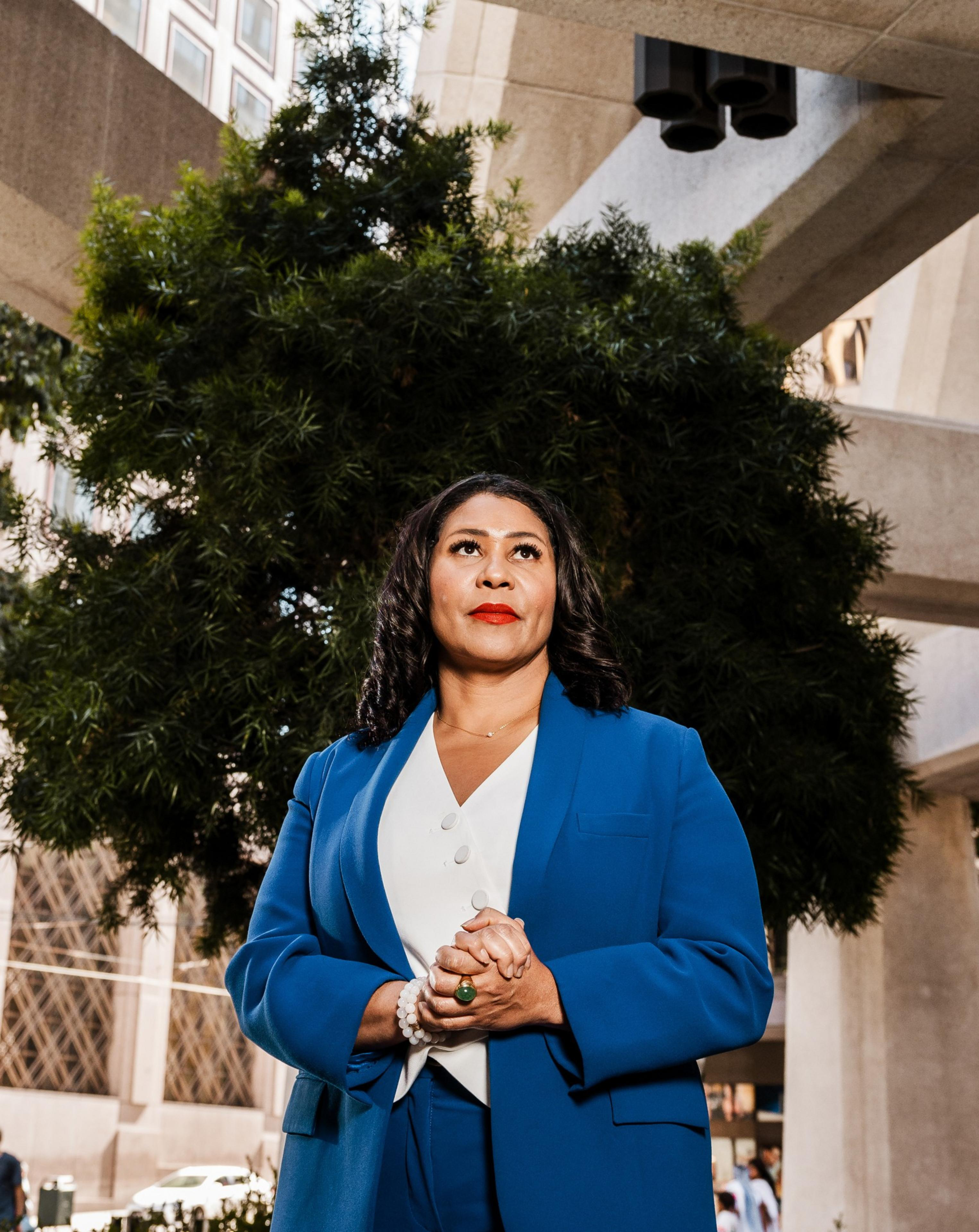 A woman stands confidently in front of a leafy green tree, wearing a blue suit and a white blouse, with her hands clasped. The background shows an urban environment.