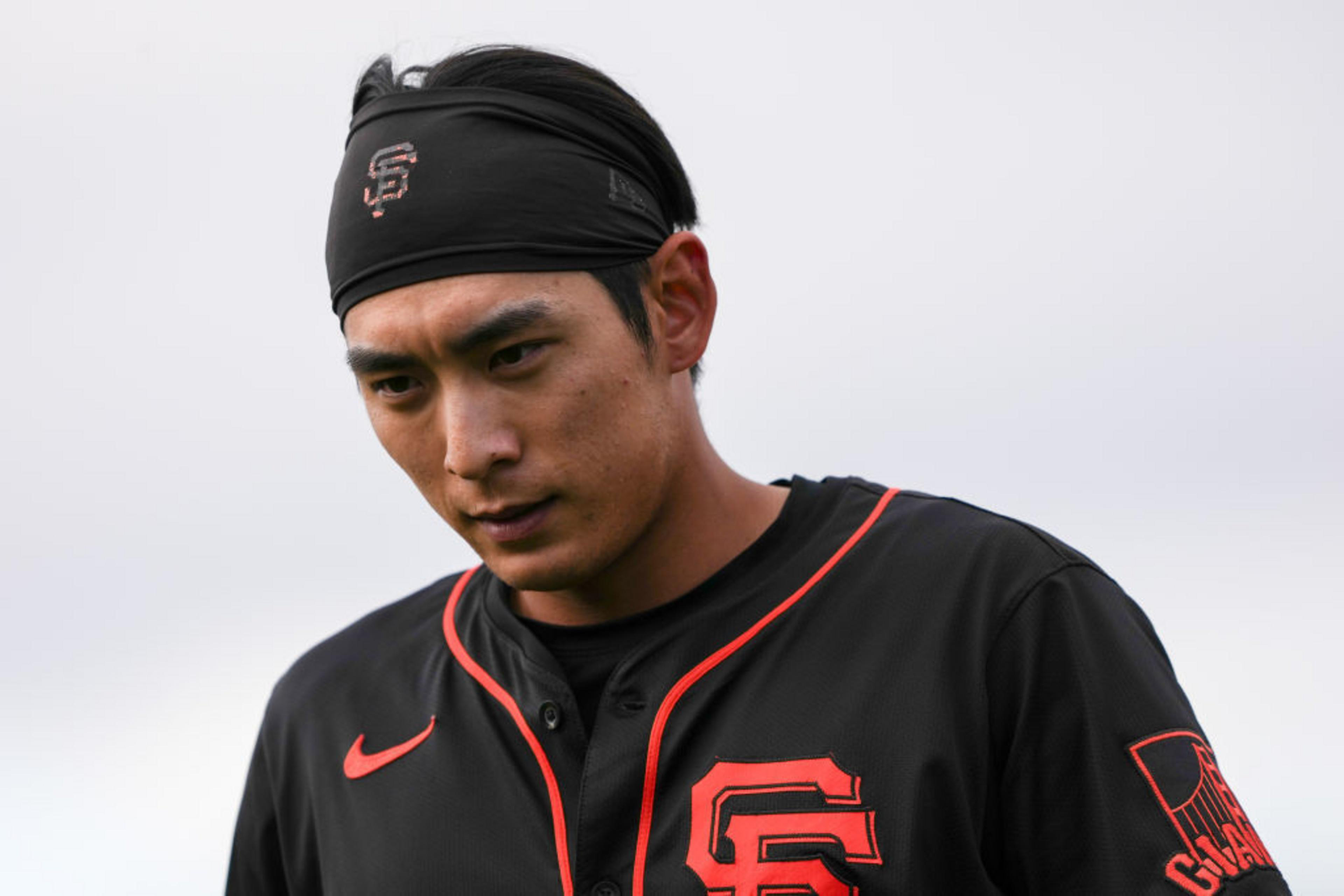 A person is wearing a black baseball jersey with orange details and a headband featuring a logo. The expression appears thoughtful or focused.