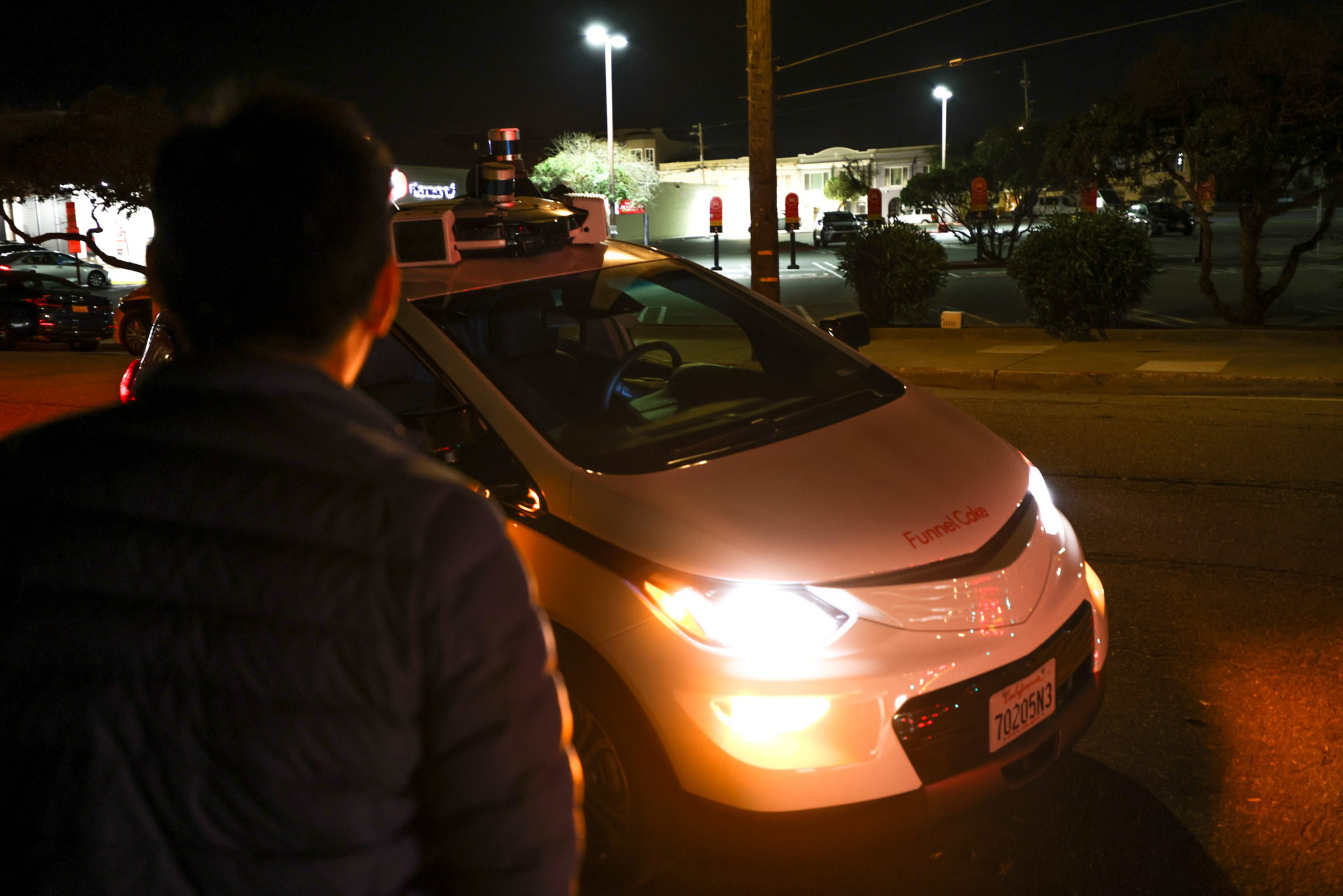A person in a dark jacket stands in front of a white autonomous car with bright headlights on a dimly lit street at night.