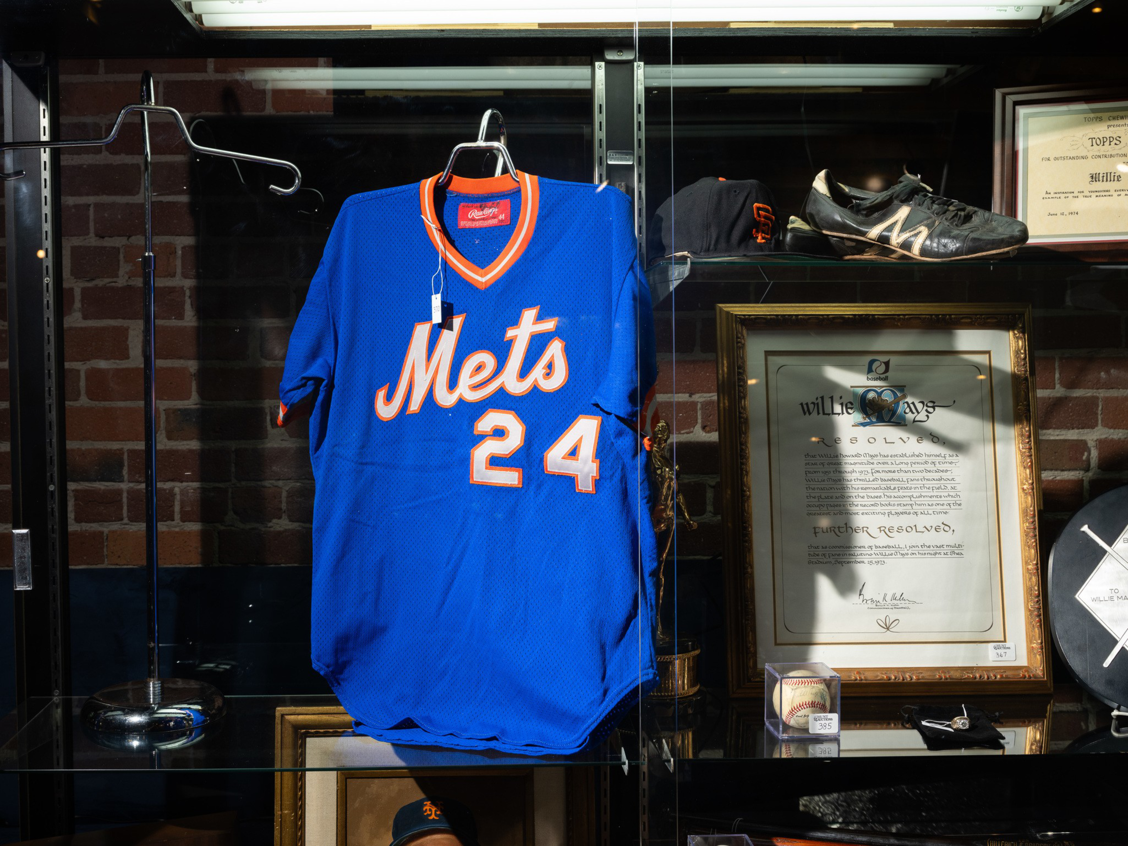 A blue Mets jersey with number 24 hangs next to a framed document, a baseball, cleats, and a black cap with an orange “SF” logo in a display case.