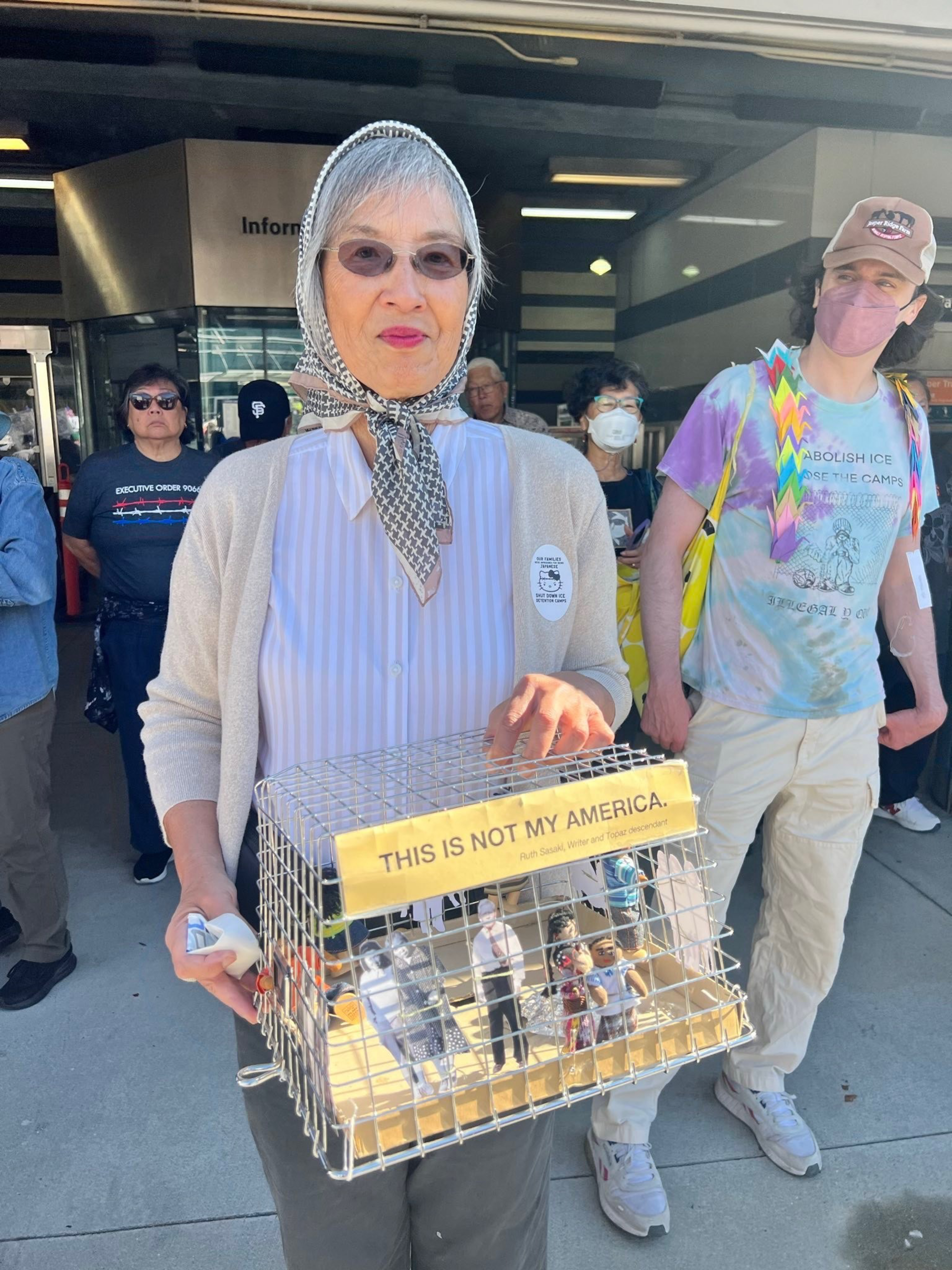 An older woman wearing a scarf and glasses holds a cage with small figures inside and a sign reading “This is not my America.”