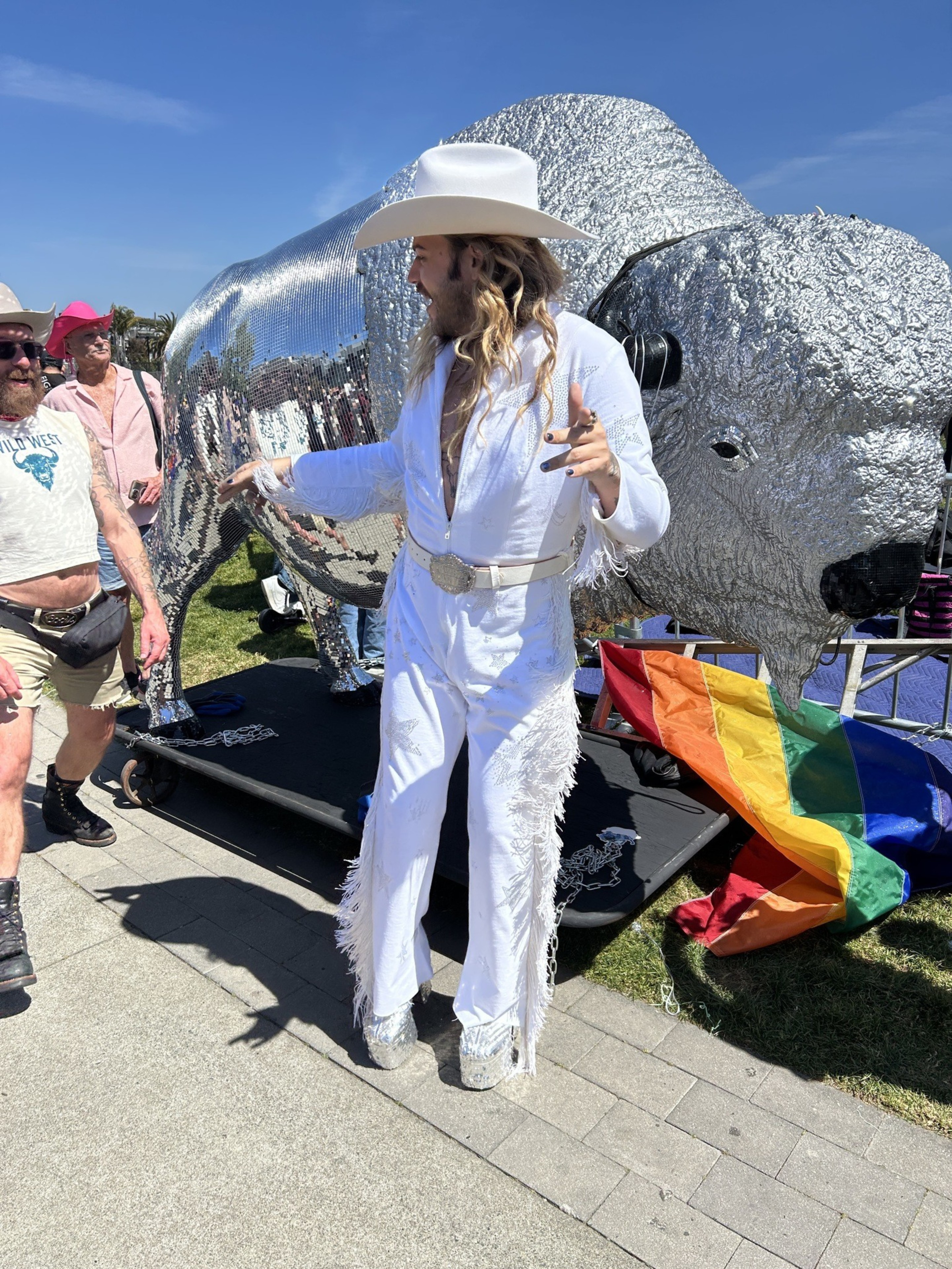 A person in a white, fringed outfit and cowboy hat stands beside a shiny, silver buffalo sculpture. A rainbow flag lies on the ground nearby.