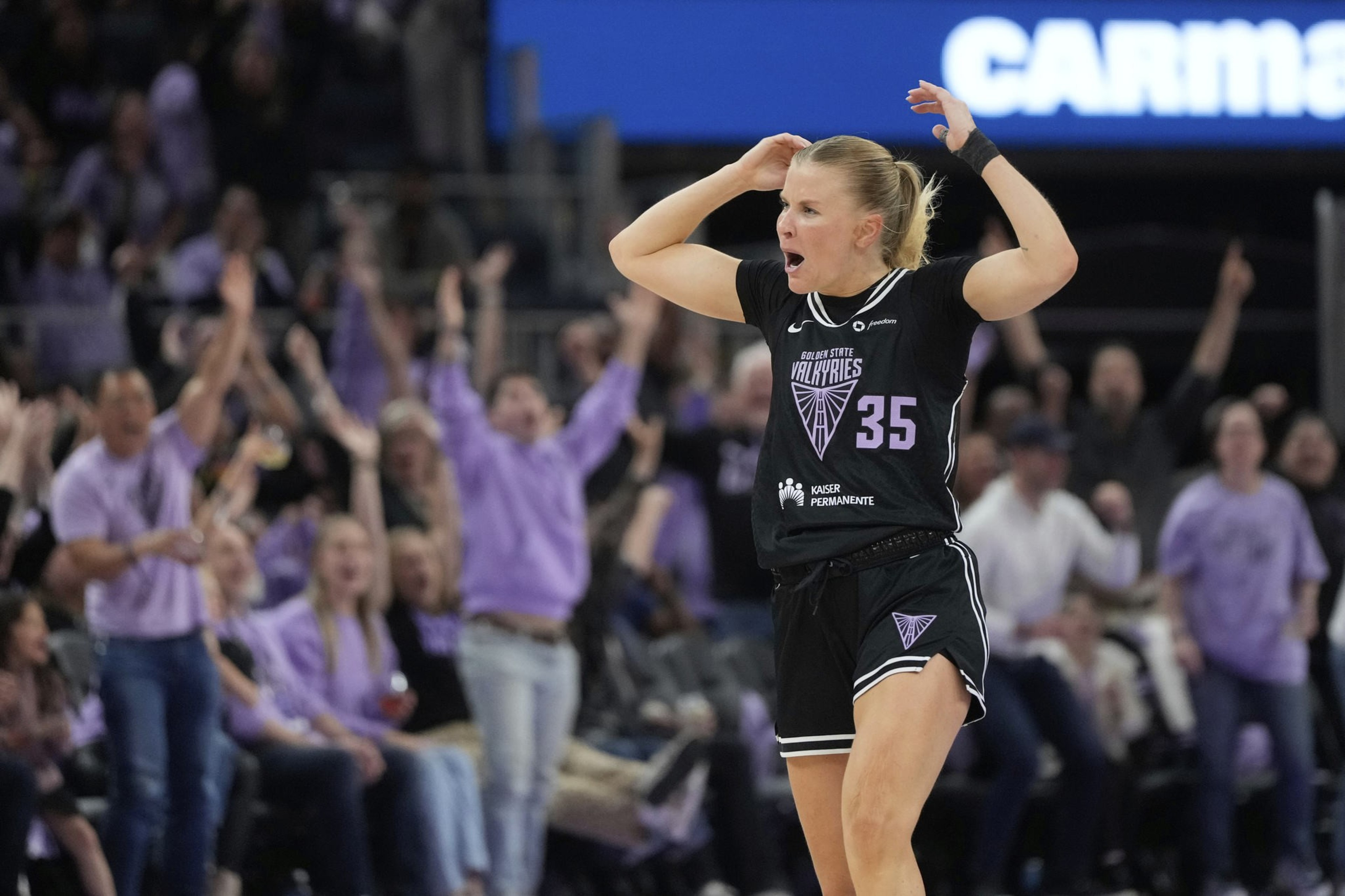 A basketball player in a black uniform gestures with excitement on the court. The crowd in the background cheers with raised arms, creating an energetic atmosphere.