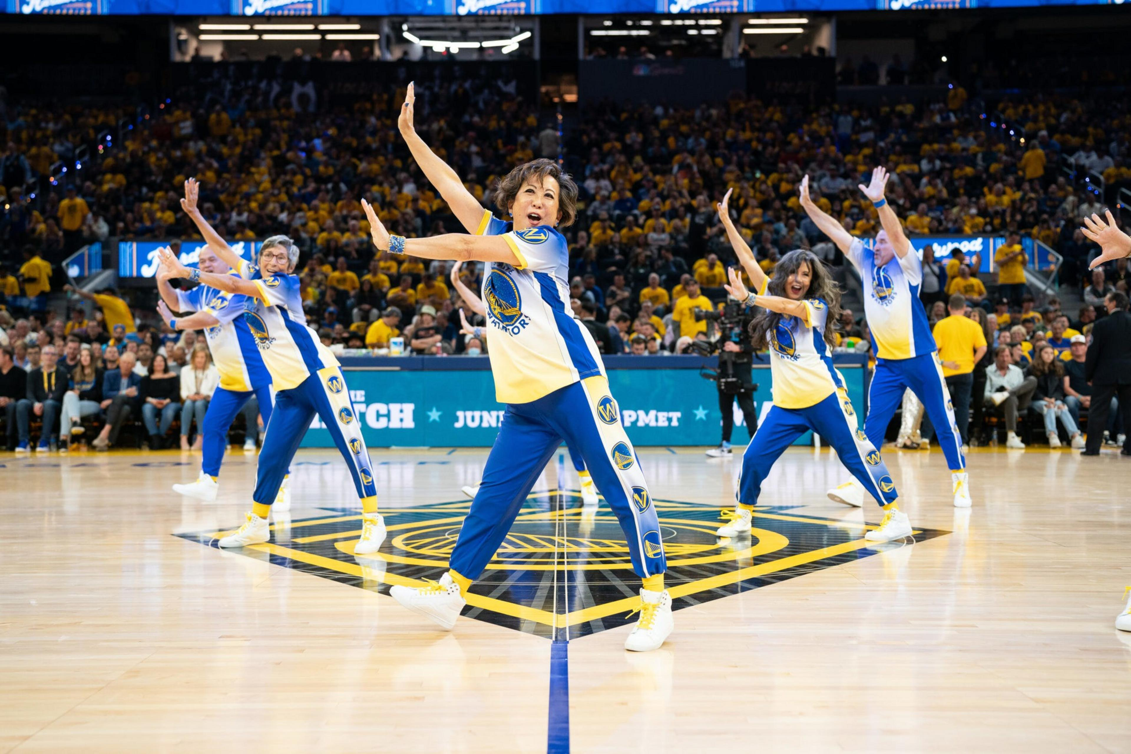 Five dancers in blue and yellow Golden State Warriors outfits perform a choreographed routine on a basketball court with a crowd watching.