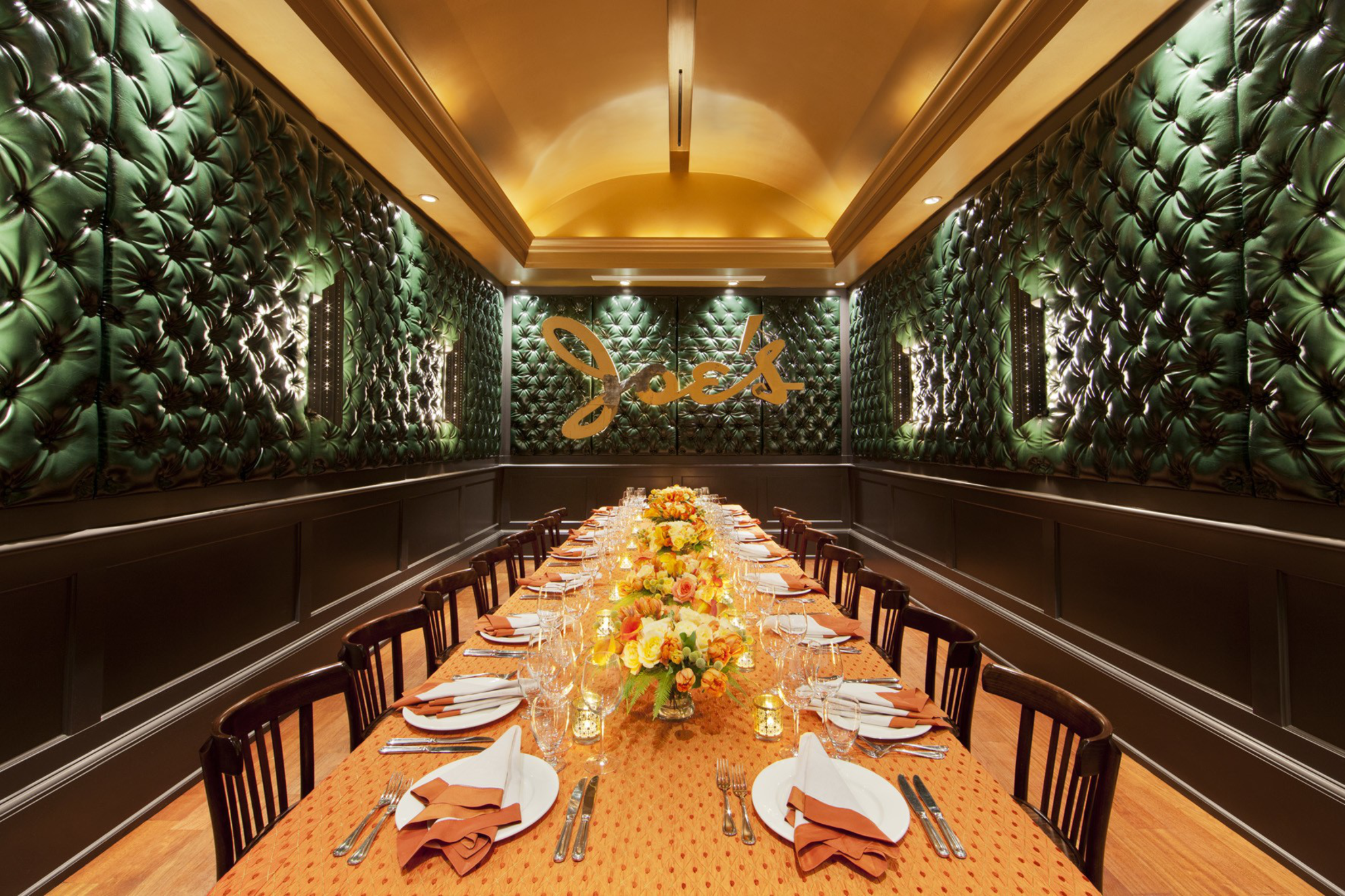 A luxurious dining room features a long table with an orange cloth, set for a meal with elegant dishware and floral centerpieces. Plush green walls add a sophisticated touch.