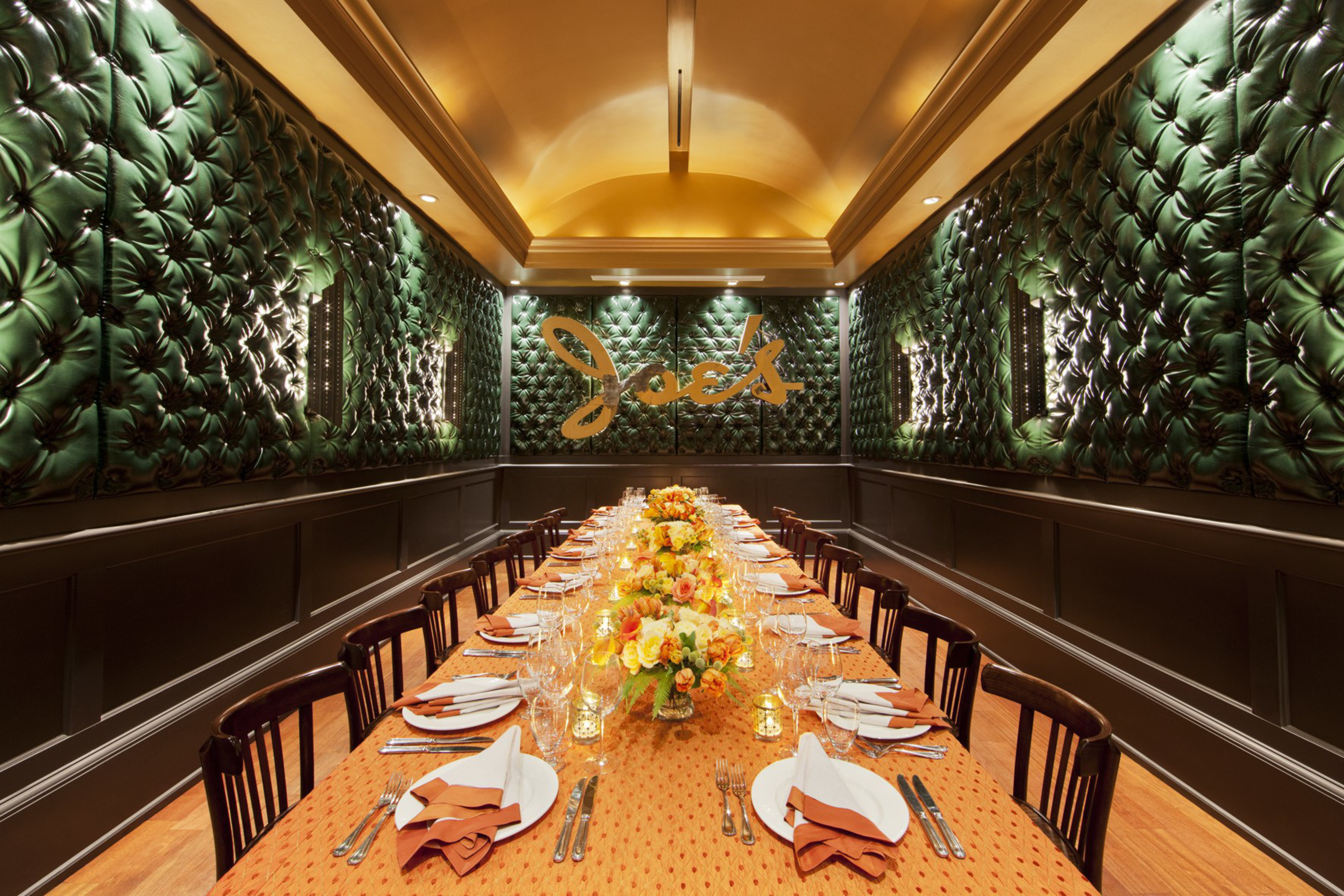 A luxurious dining room features a long table with an orange cloth, set for a meal with elegant dishware and floral centerpieces. Plush green walls add a sophisticated touch.