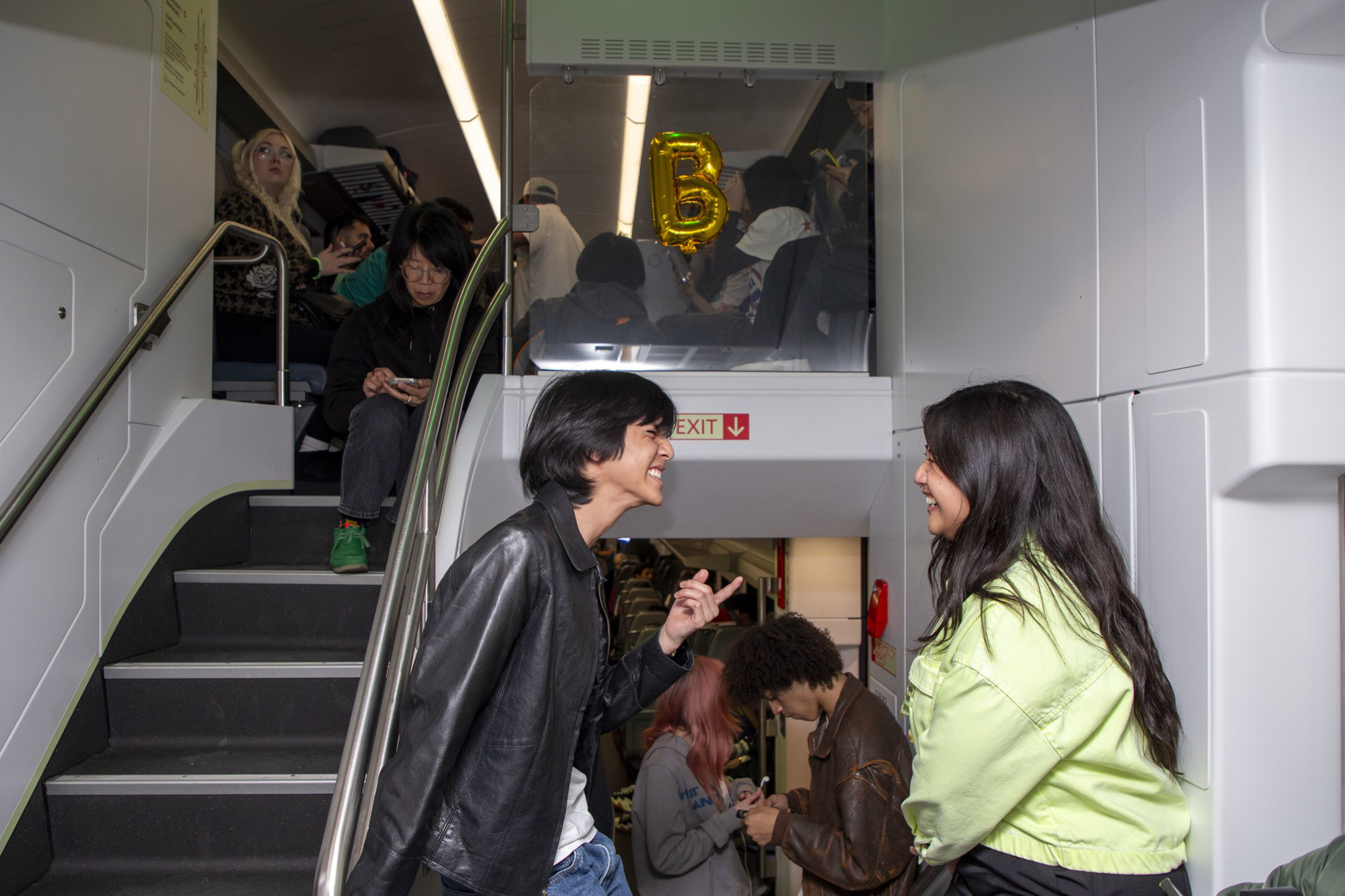Several people are on a train, chatting and using phones. A staircase leads to an upper level where a gold “B” balloon is visible.